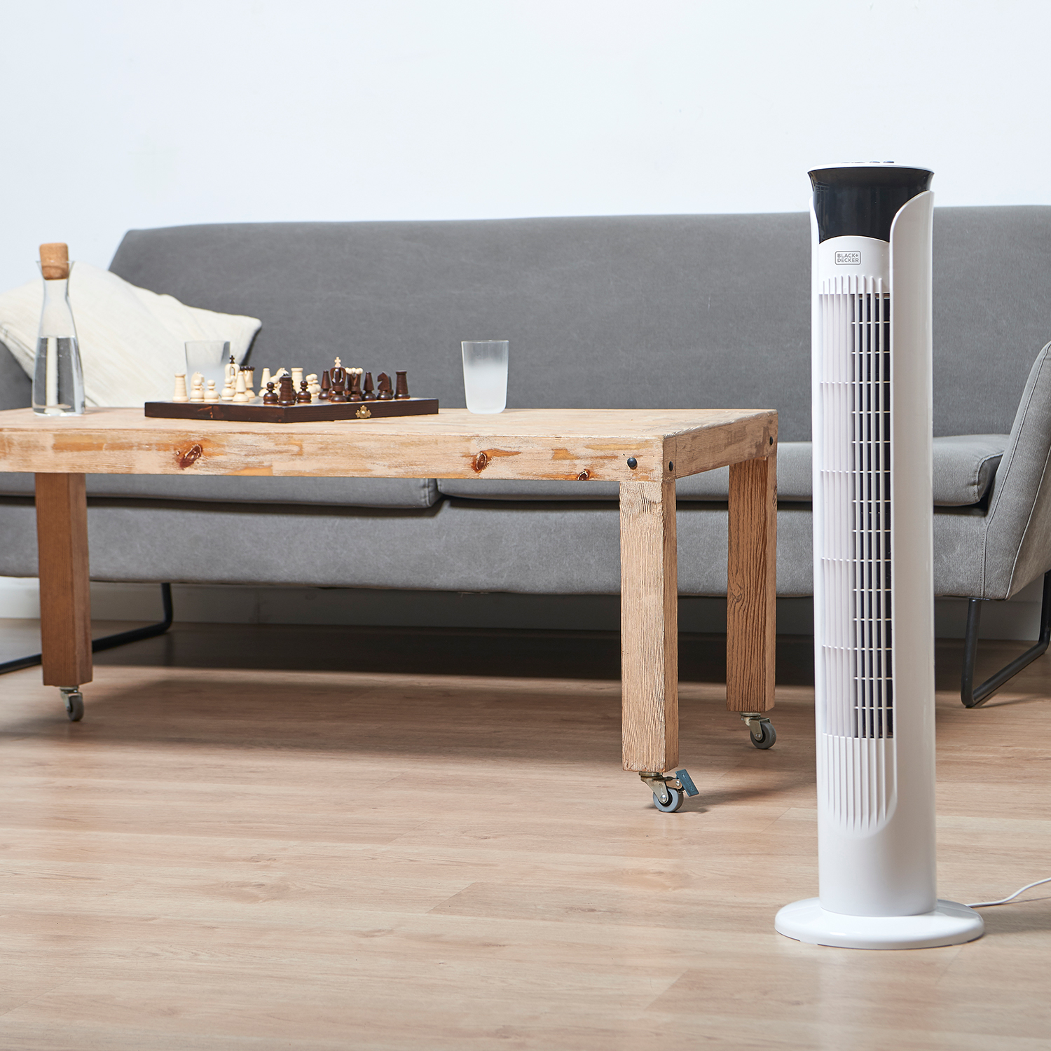 Weißer Turmventilator, Holztisch mit Schach, Sofa im Hintergrund. Heller Raum.