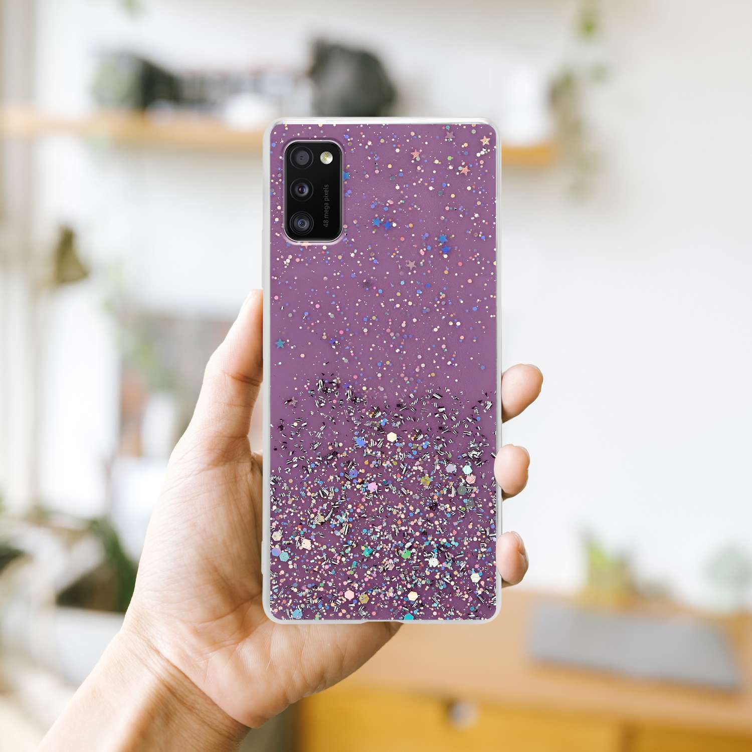 Eine Hand, die eine lila Handyhülle mit Glitzer hält, mit verschwommenem Hintergrund eines Zimmers.