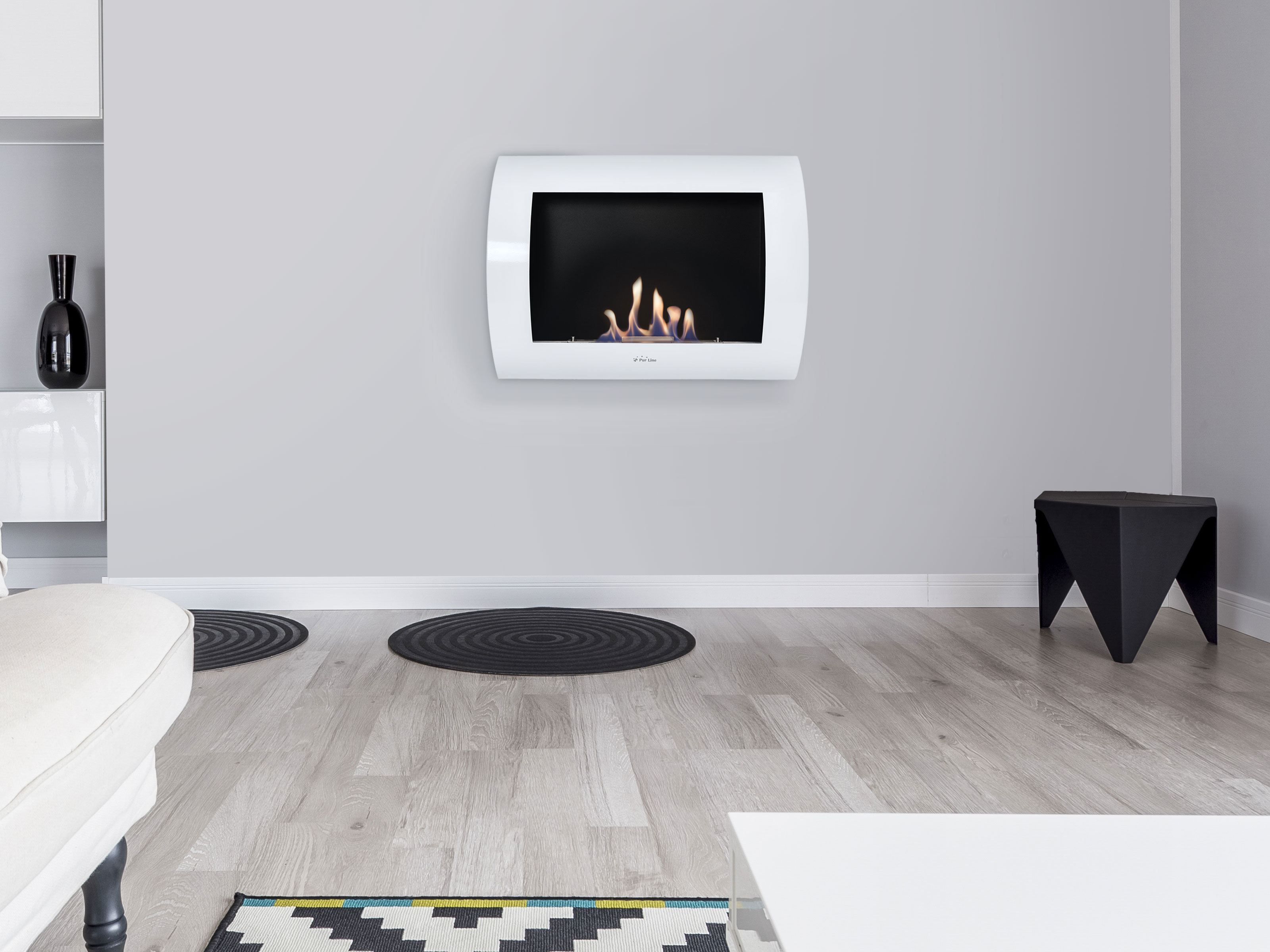 Ein weißer, an der Wand befestigter Kamin mit Flammen. Eine schwarze Vase und ein Hocker sind in der Nähe.