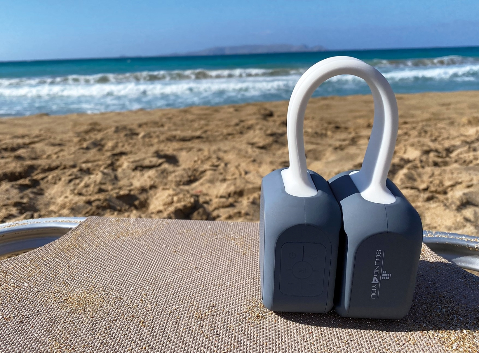 Zwei graue Lautsprecher mit weißem Griff am Strand. Wellen im Hintergrund.