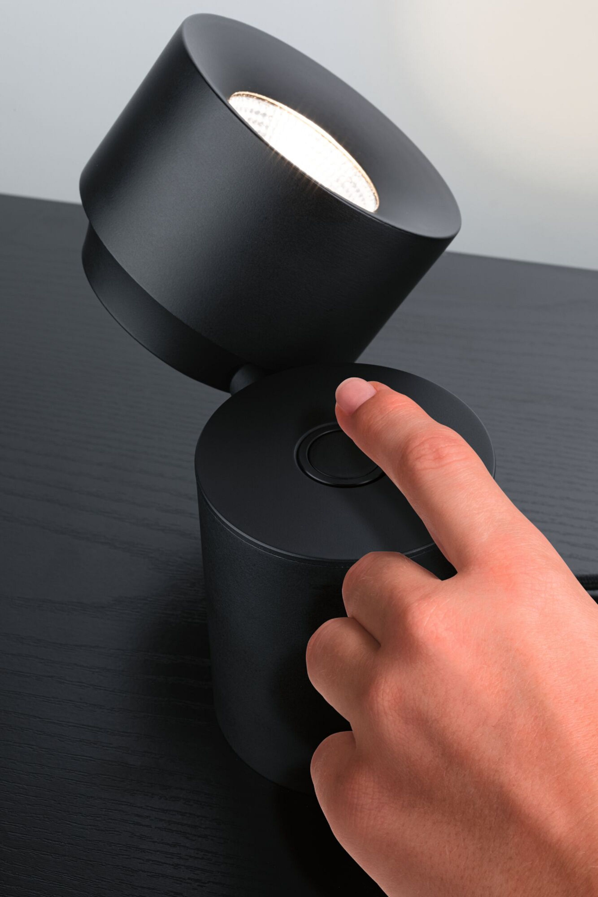 Eine schwarze Lampe auf einer dunklen Oberfläche. Ein Finger drückt einen Knopf am Sockel der Lampe.