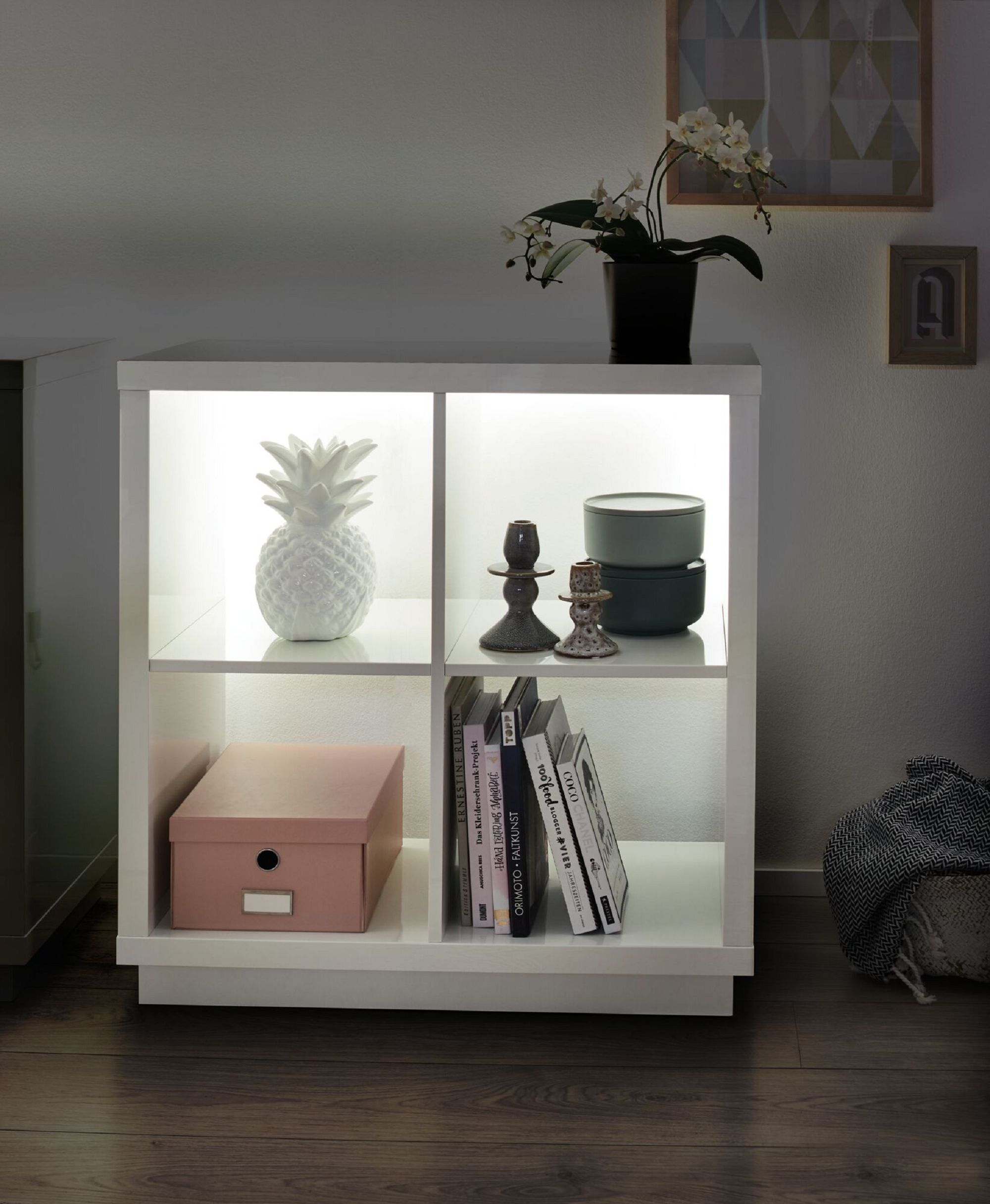 Ein weißes Würfelregal mit Gegenständen: Ananas, Kerzen, Bücher und eine rosa Box.