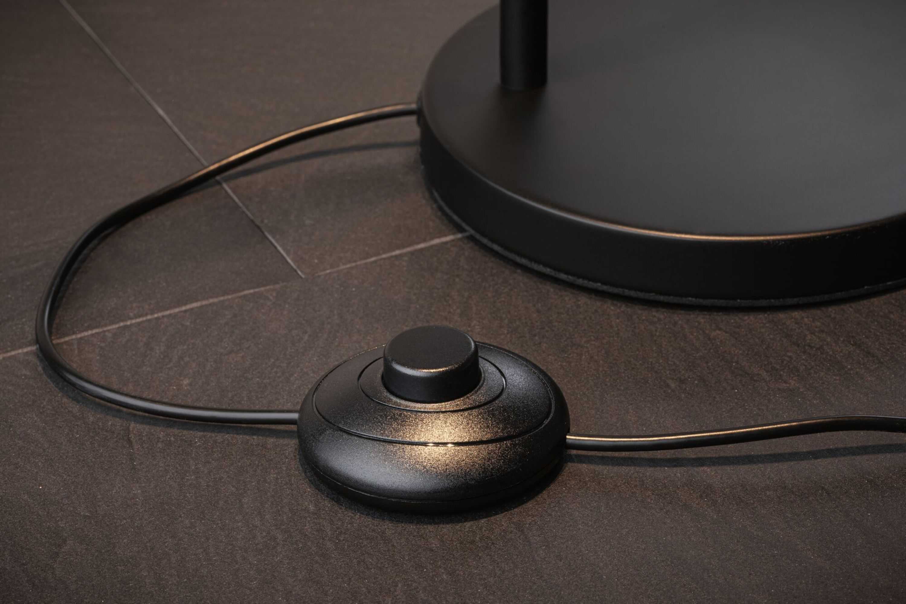 Czarny przełącznik ściemniacza nożnego znajduje się na ciemnej kamiennej podłodze z czarnym kablem.