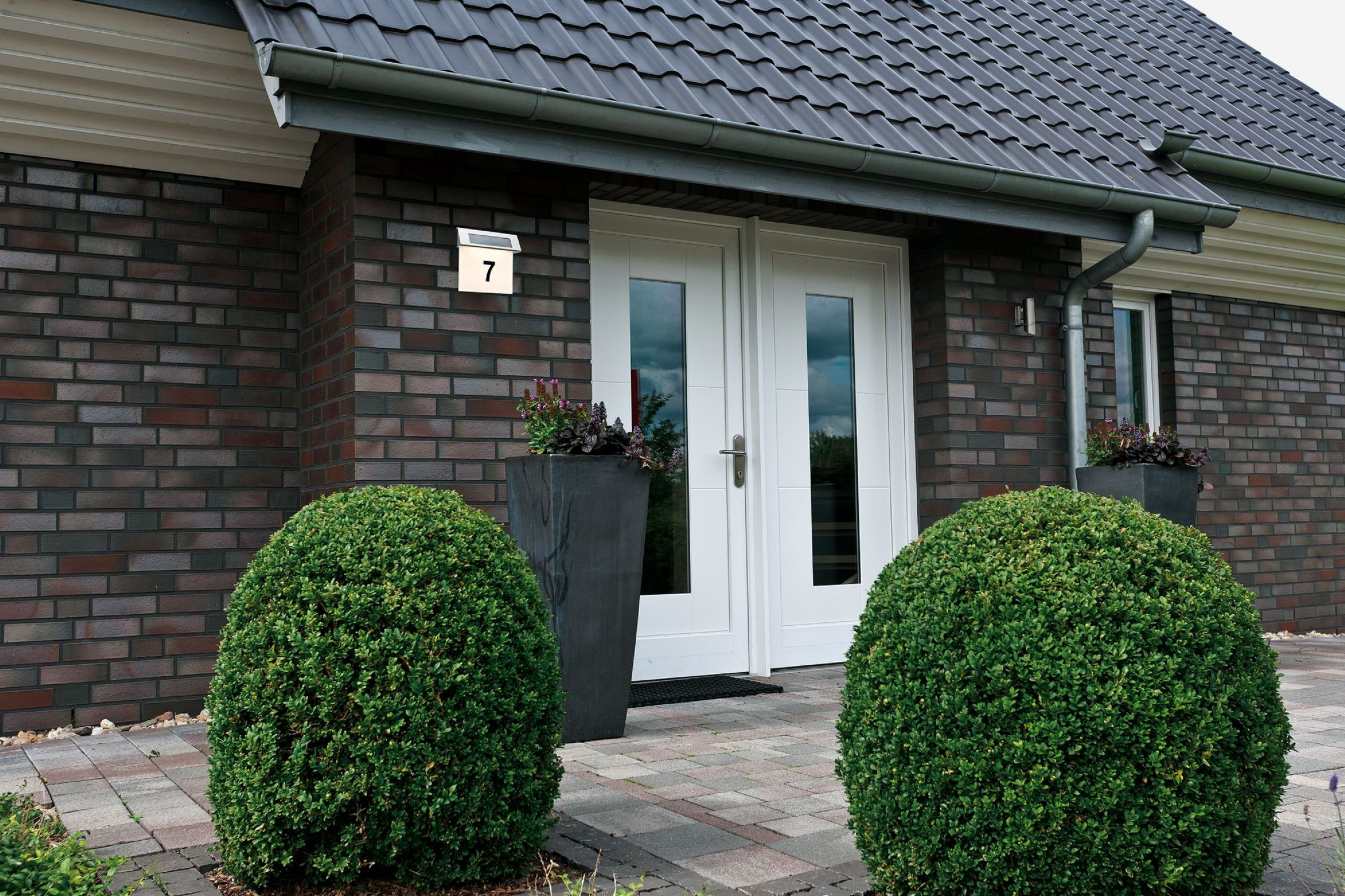 Elewacja domu z cegieł, podwójnymi drzwiami i zielonymi krzewami. Numer 7 na ścianie. Dach z płytek.