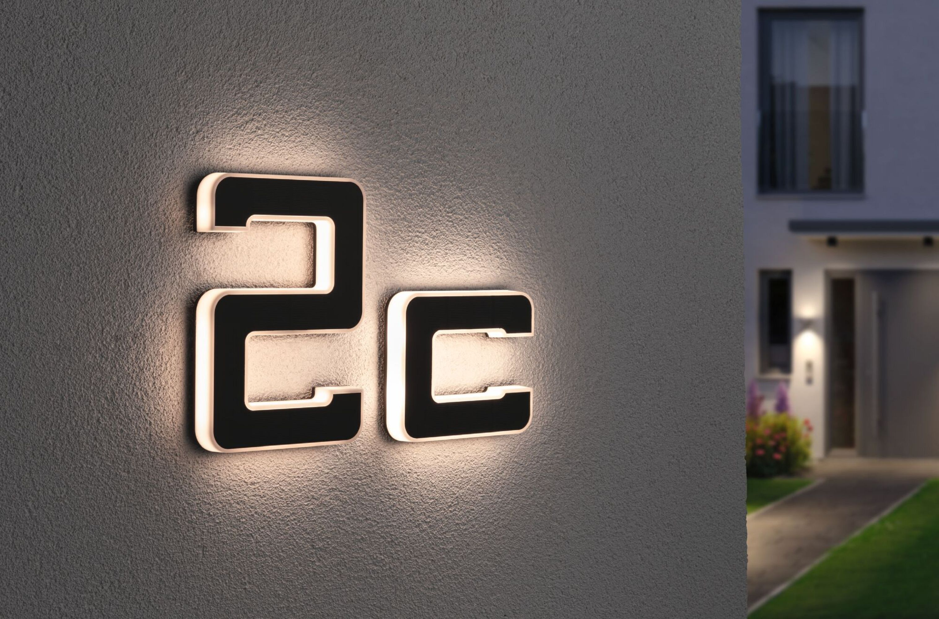Beleuchtete Hausnummern '2C' an einer grauen Wand, mit verschwommenem Haus und Nachthimmel im Hintergrund.