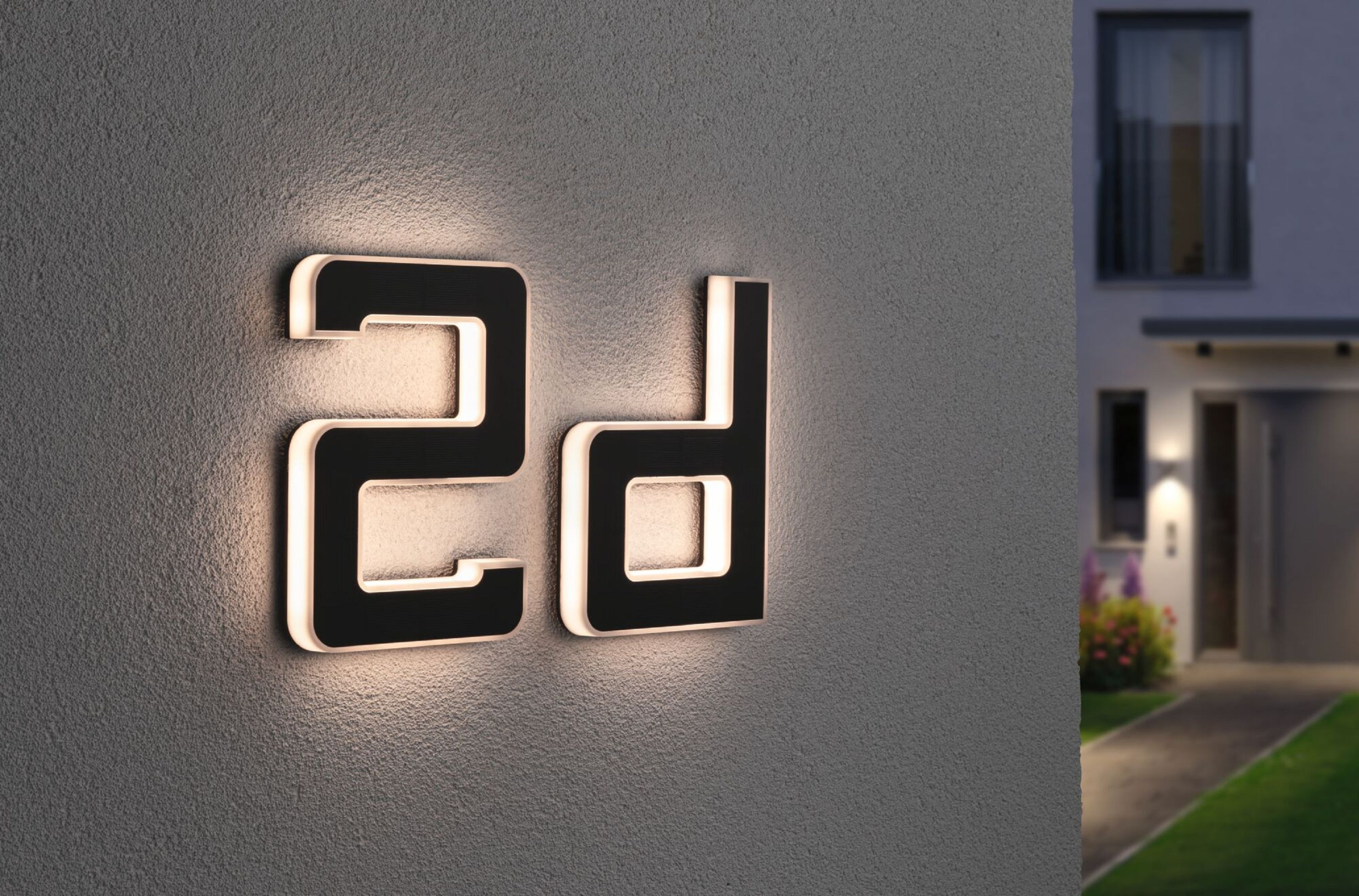 Czarny podświetlany znak z literami '2d' na szarej ścianie. Dom i ścieżka w tle.