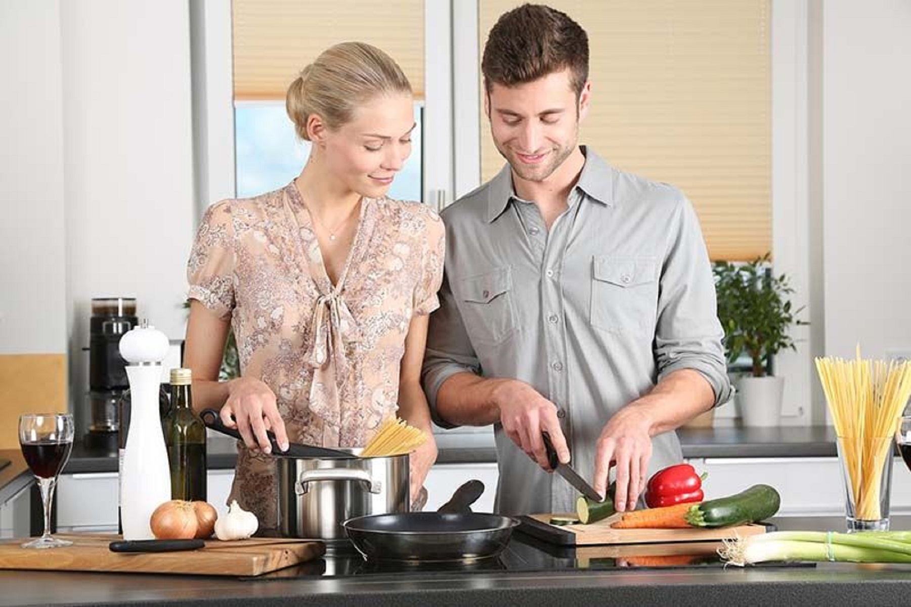 Una pareja cocina en una cocina. La mujer agrega pasta a una olla. Un hombre corta verduras en una tabla de cortar.