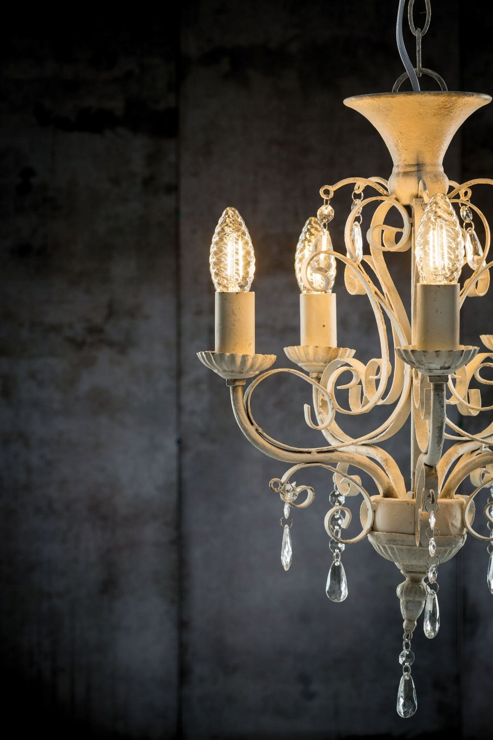 Biały żyrandol z trzema zapalonymi żarówkami, kryształami i ozdobnym wzorem, na tle ciemnej ściany.