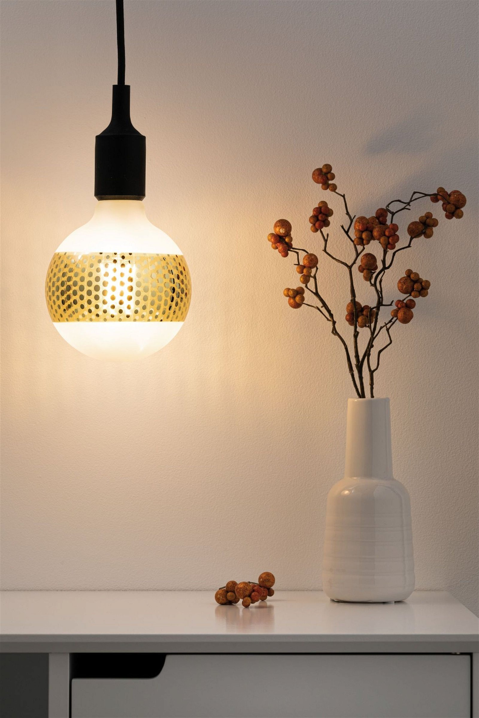 Lampadina decorativa con pois dorati, illuminata, con vaso di bacche su tavolo.