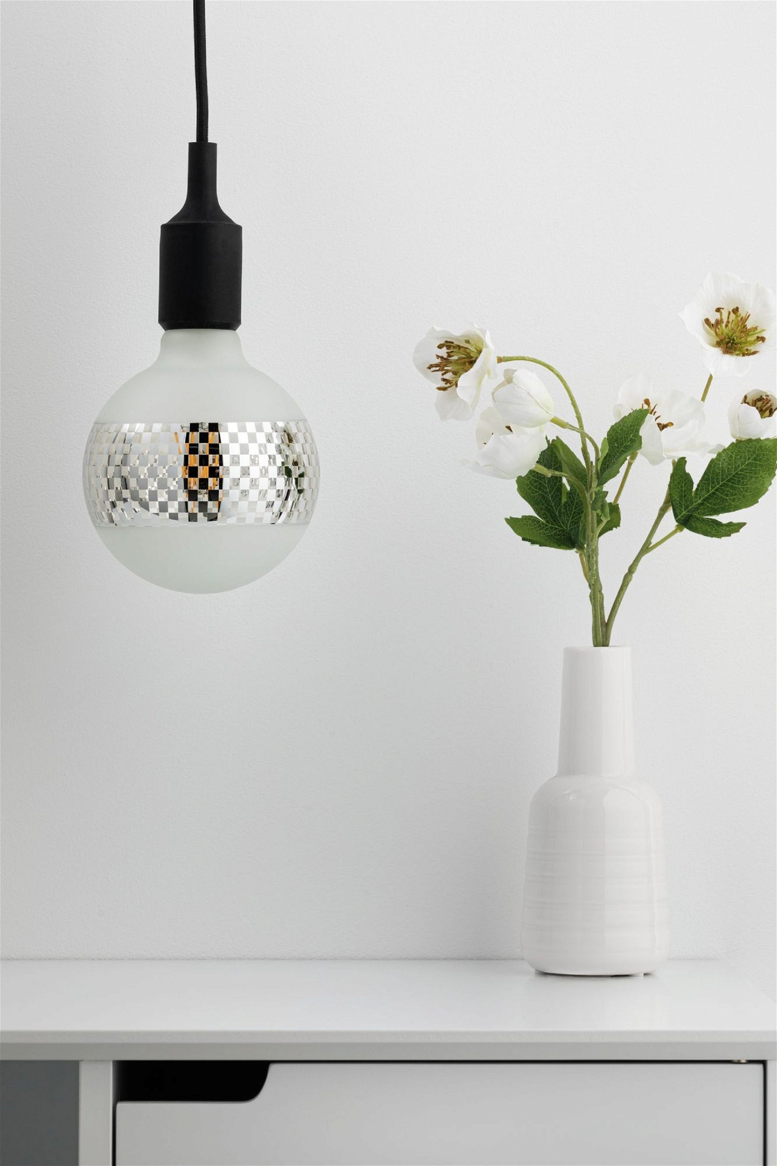 Eine moderne Glühbirne mit Schachbrettmuster hängt, und rechts steht eine Vase mit Blumen.