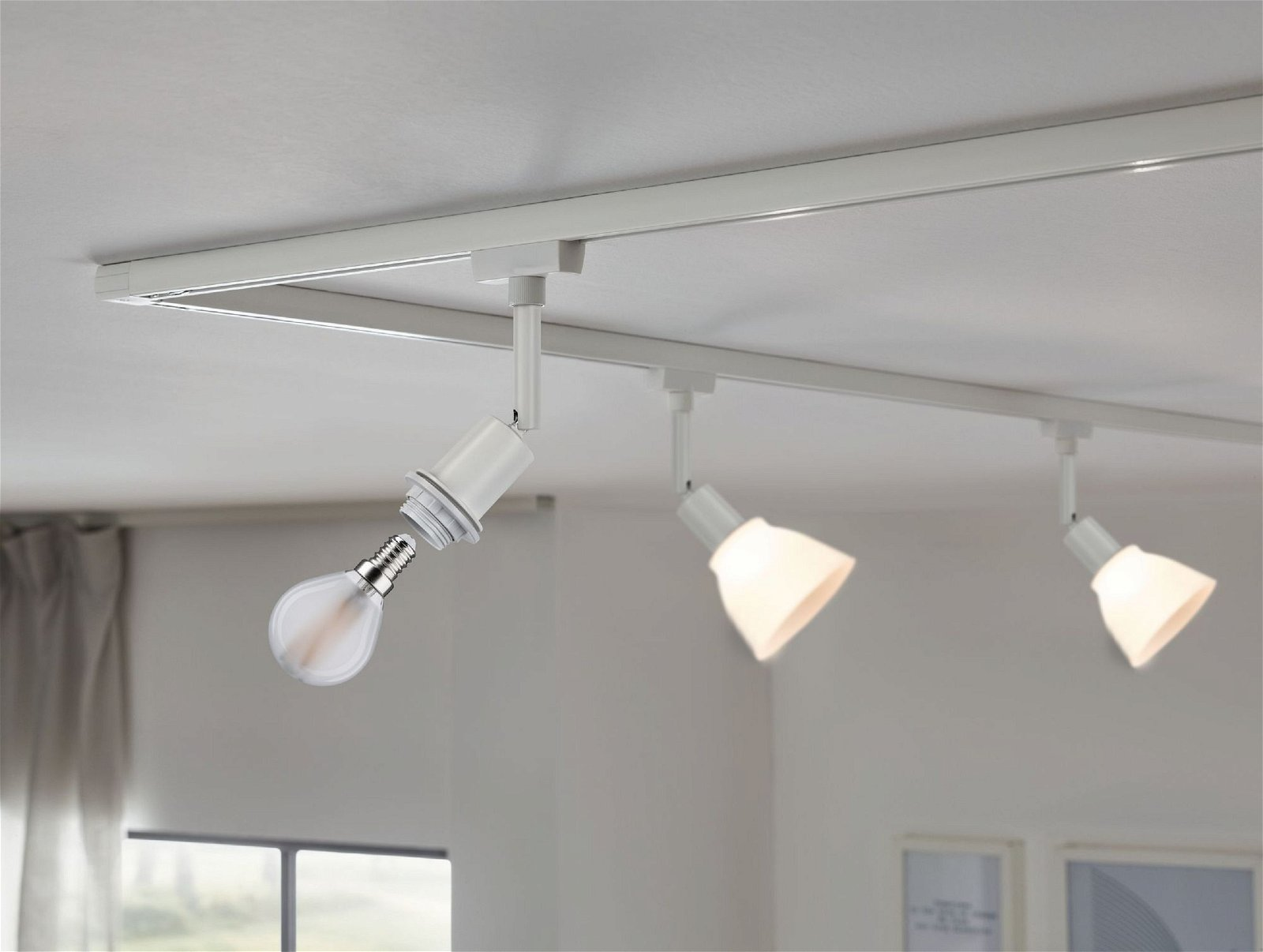 Schienenbeleuchtung mit drei Strahlern, einer mit freiliegender Glühbirne. Weiße Decke und Raum.