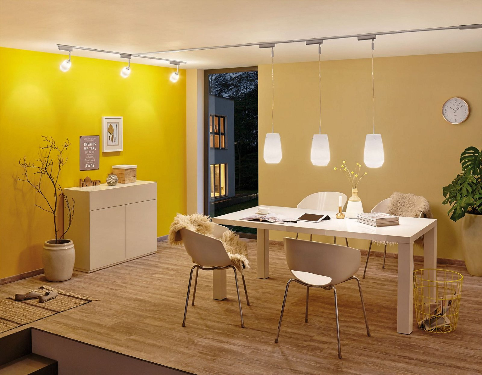 Jadalnia z żółtymi ścianami, białym stołem, krzesłami i wiszącymi lampami. Okno pokazuje widok na zewnątrz.