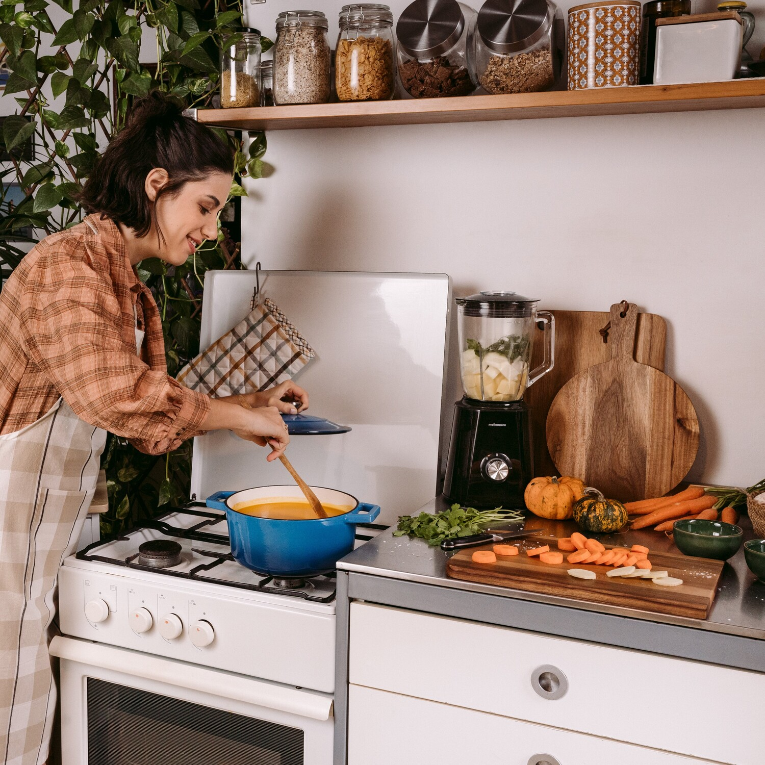 Mujer cocinando con una olla azul en la estufa, rodeada de ingredientes y utensilios.