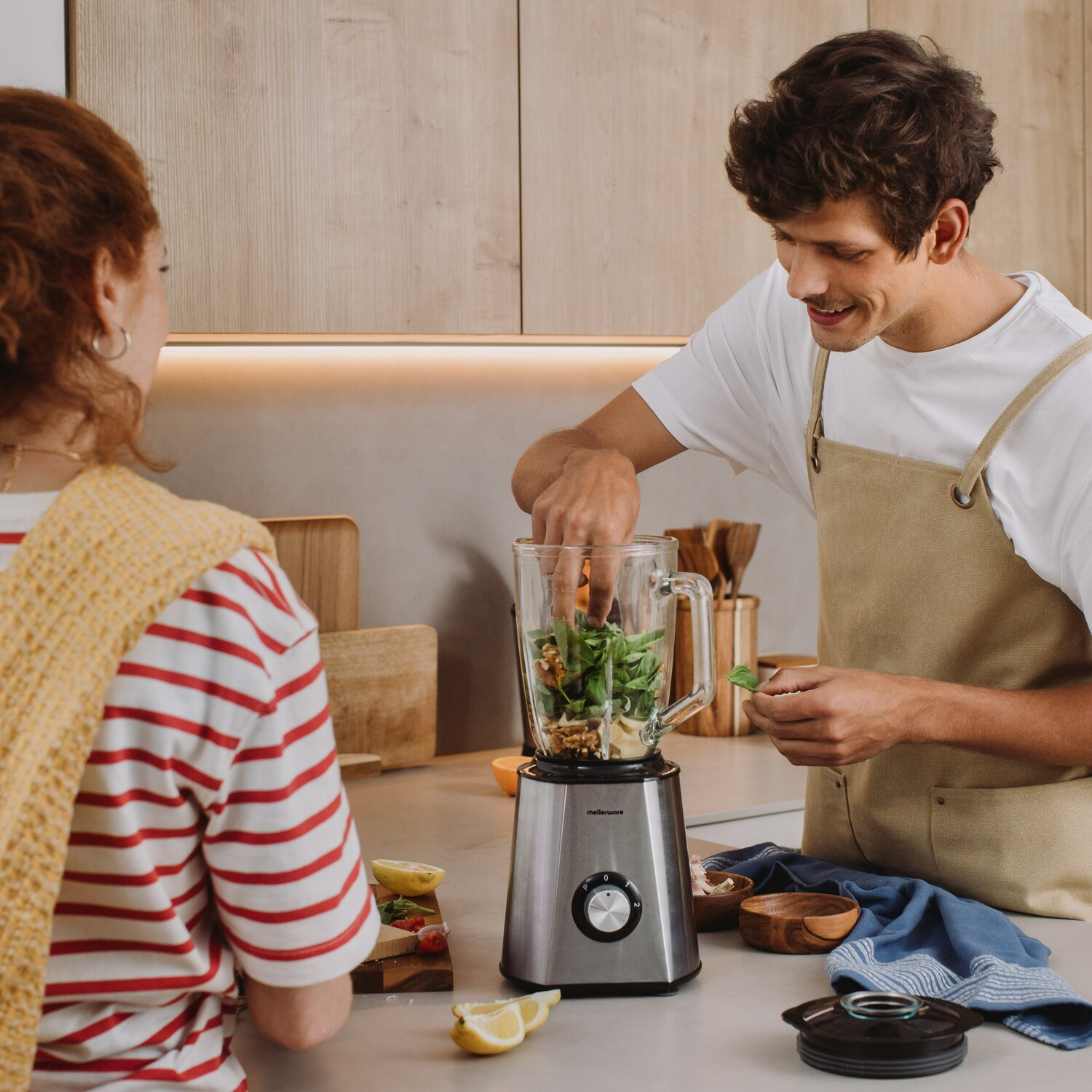 Hombre y mujer usando una licuadora, ingredientes visibles, ambiente de cocina de madera.