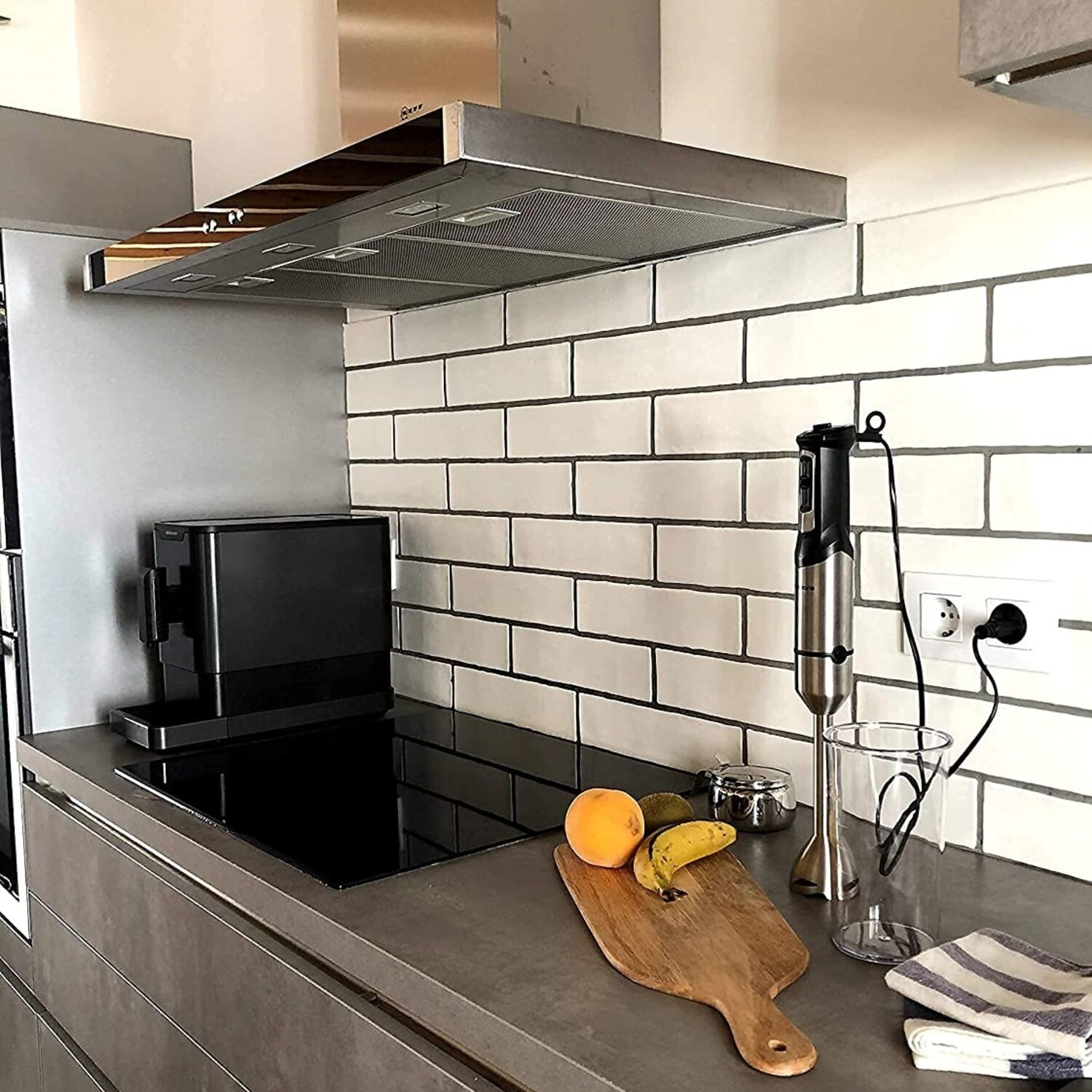 Una scena di cucina con un piano cottura nero e una cappa in acciaio inox. Sono presenti un frullatore e frutta.
