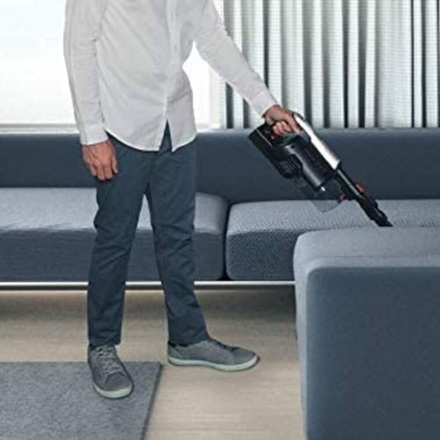 Un uomo aspira un divano blu con un aspirapolvere portatile. Indossa una camicia bianca e pantaloni scuri.