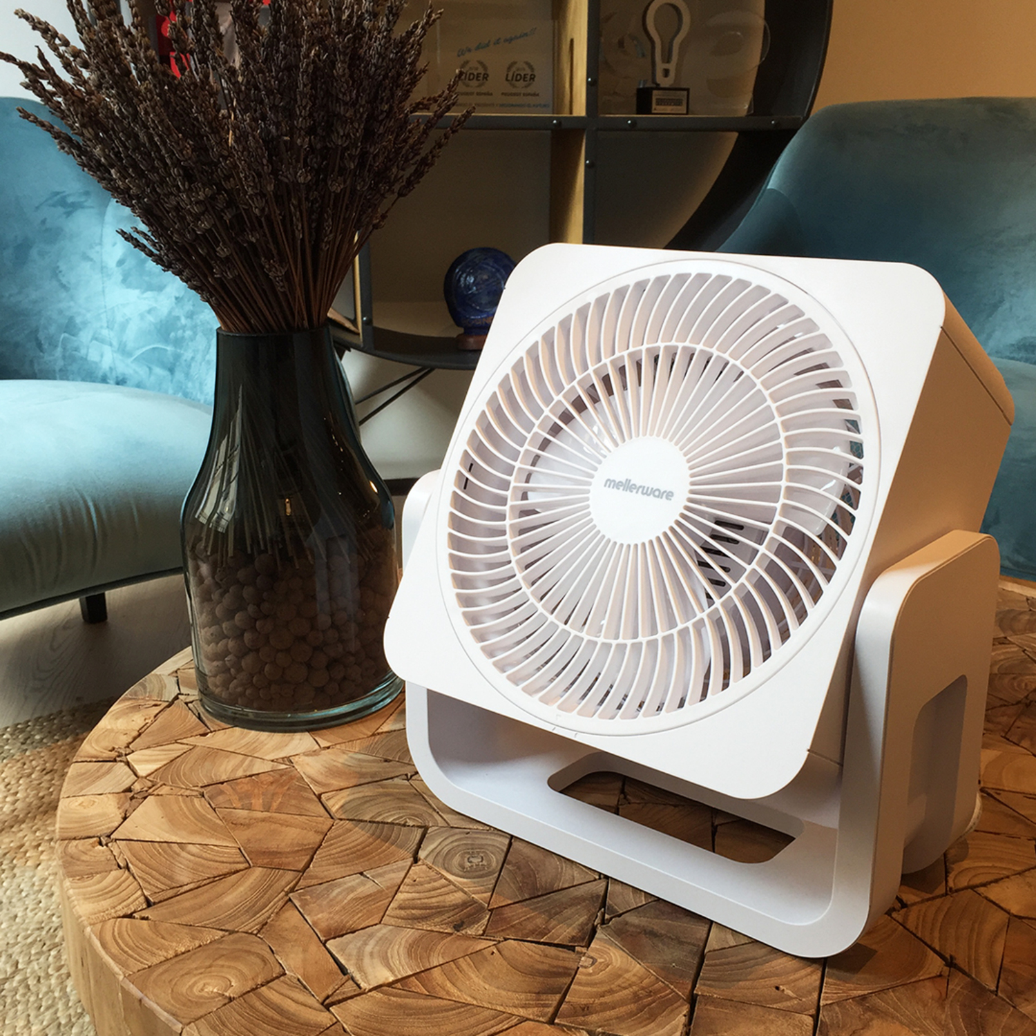 Ventilatore bianco su un tavolo di legno, con un vaso e sedie blu sullo sfondo.