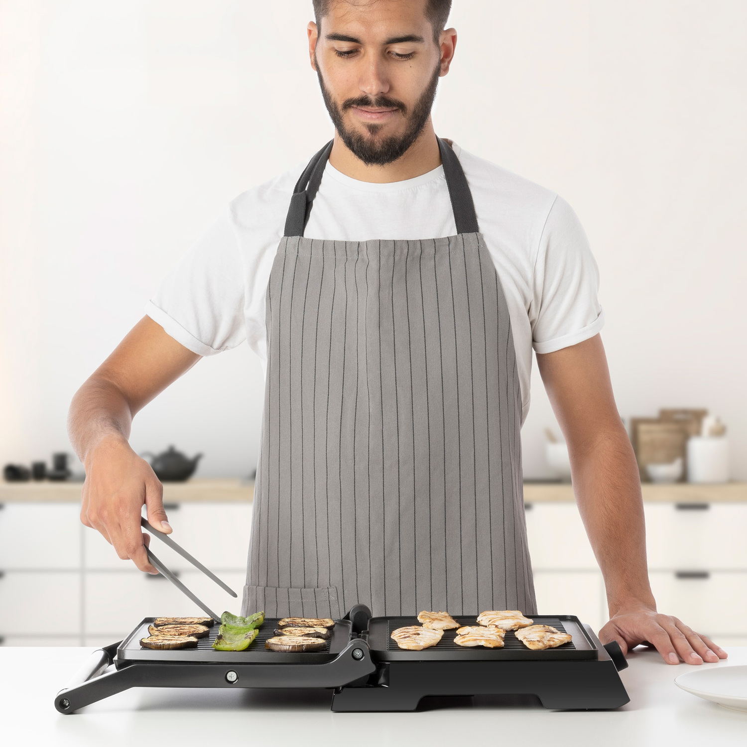 Un hombre asando verduras en una parrilla negra con delantal, con fondo blanco.