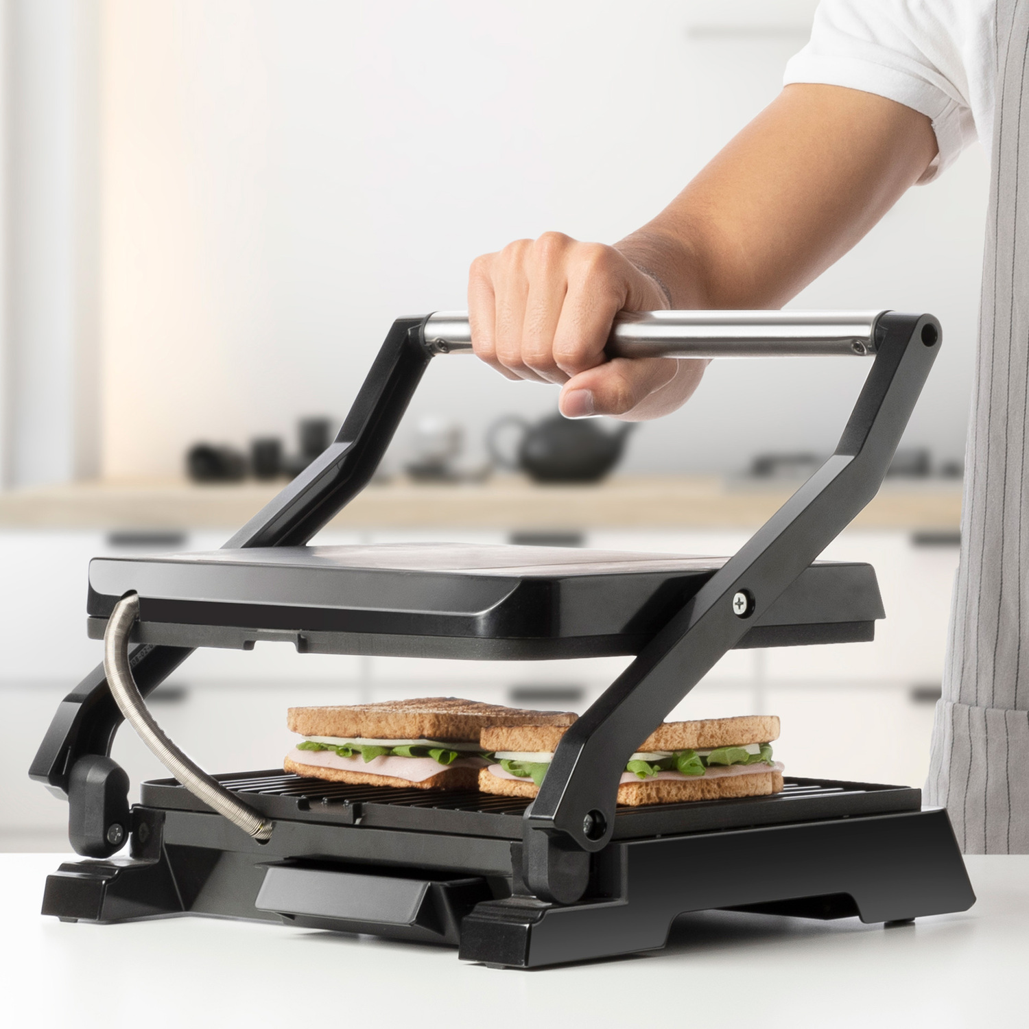 Persona presionando una sandwichera. Dos sándwiches dentro, la máquina es negra. Fondo blanco.