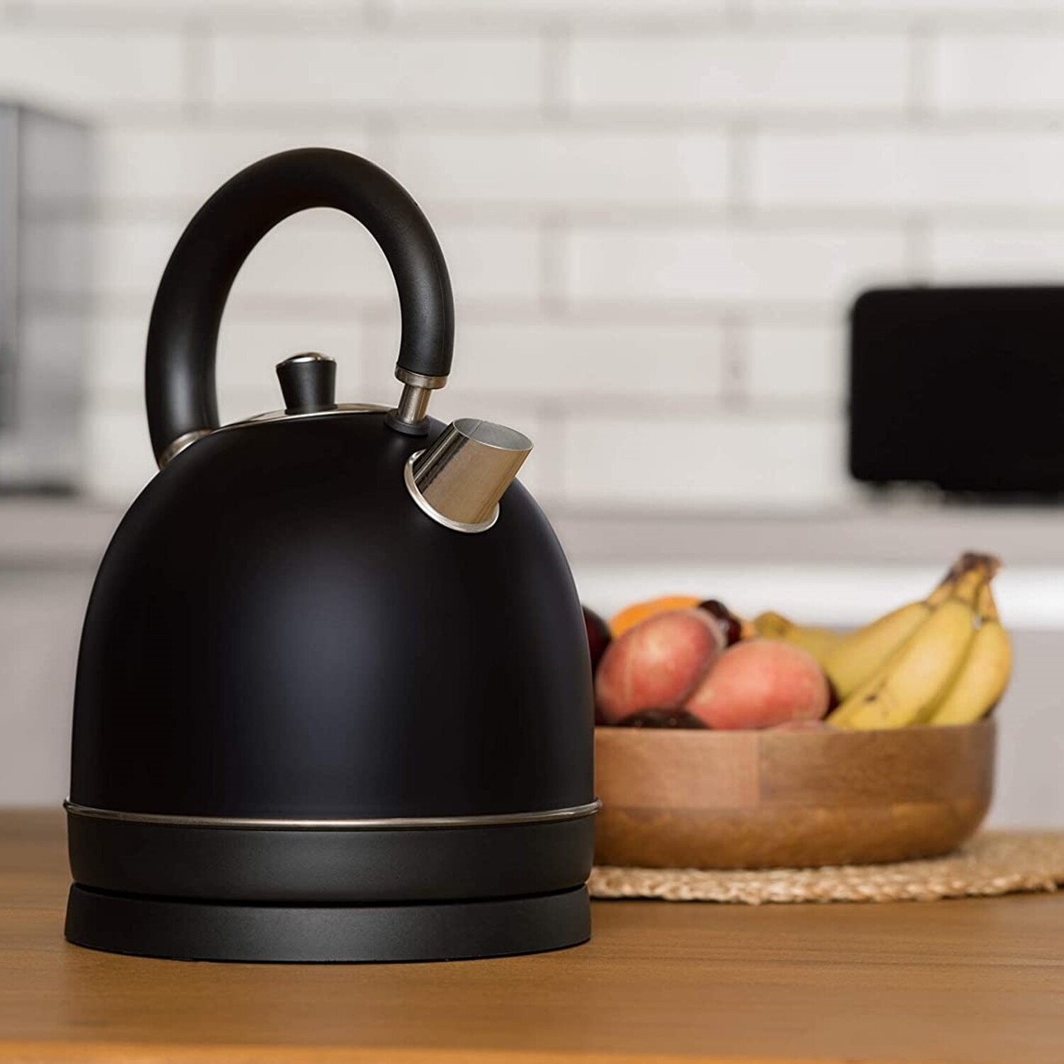 Hervidor negro en base negra, con frutero sobre mesa de madera frente a pared de ladrillo.