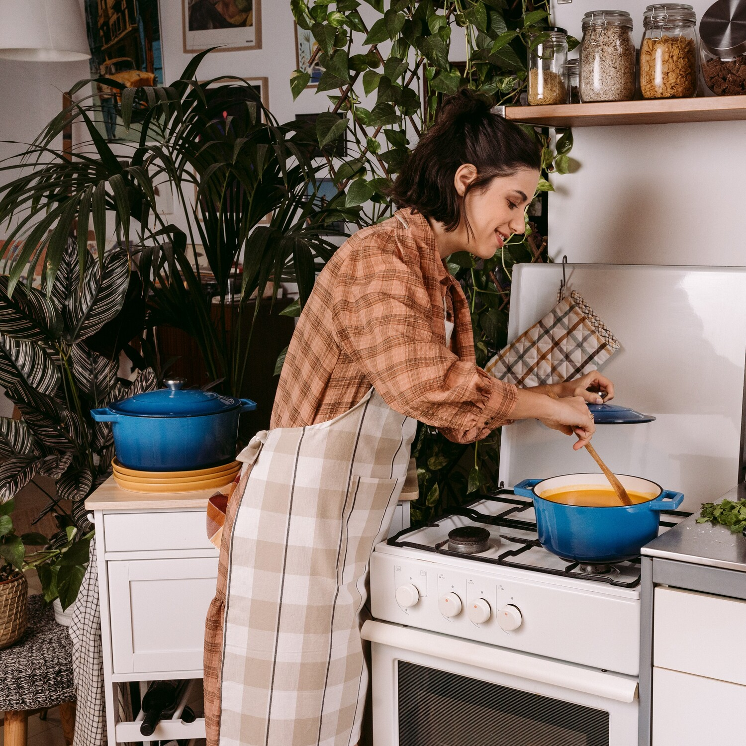 Mujer en la cocina cocinando en olla azul en la estufa, rodeada de plantas. Utensilios.