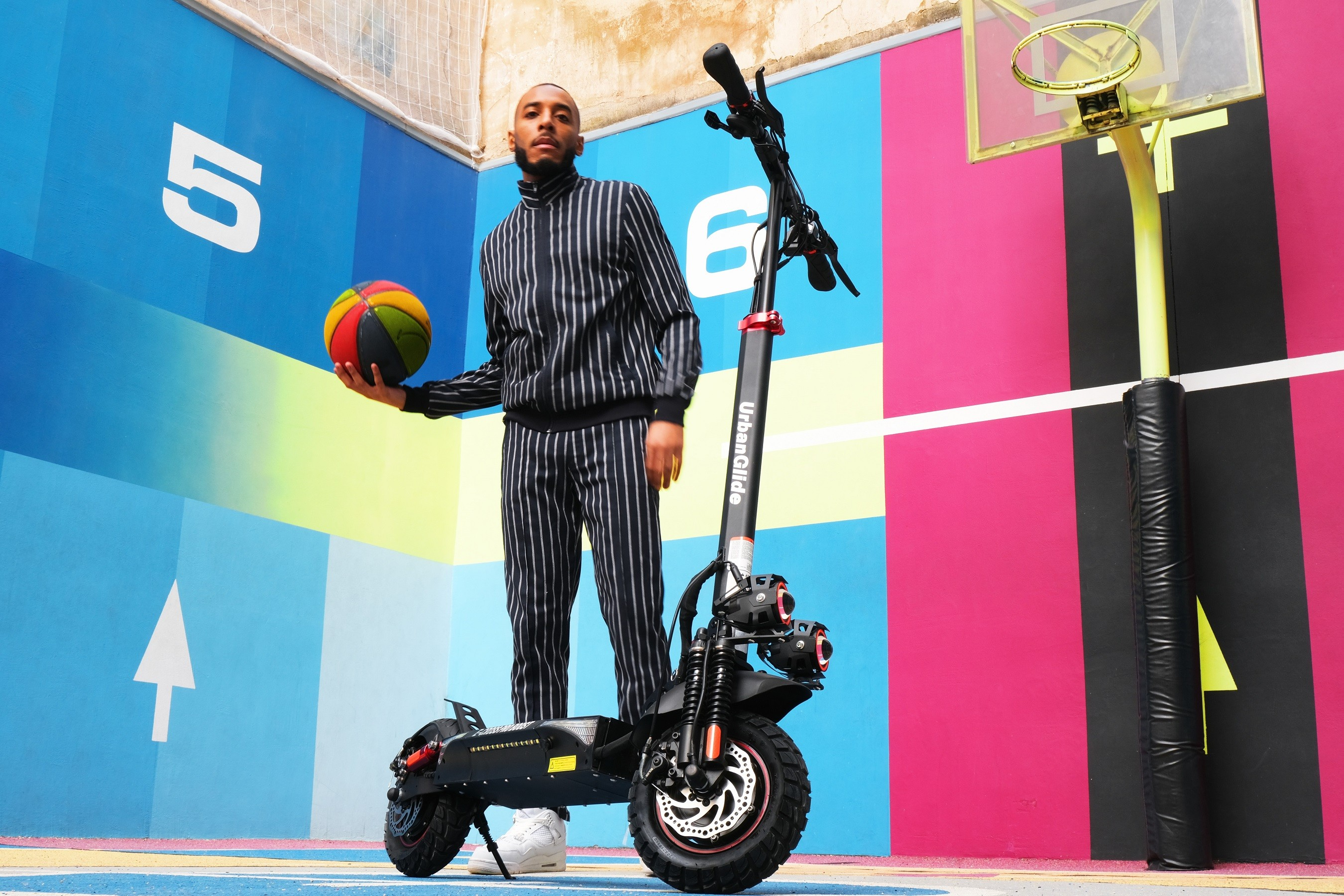 Un hombre con balón de baloncesto y patinete en una cancha colorida.