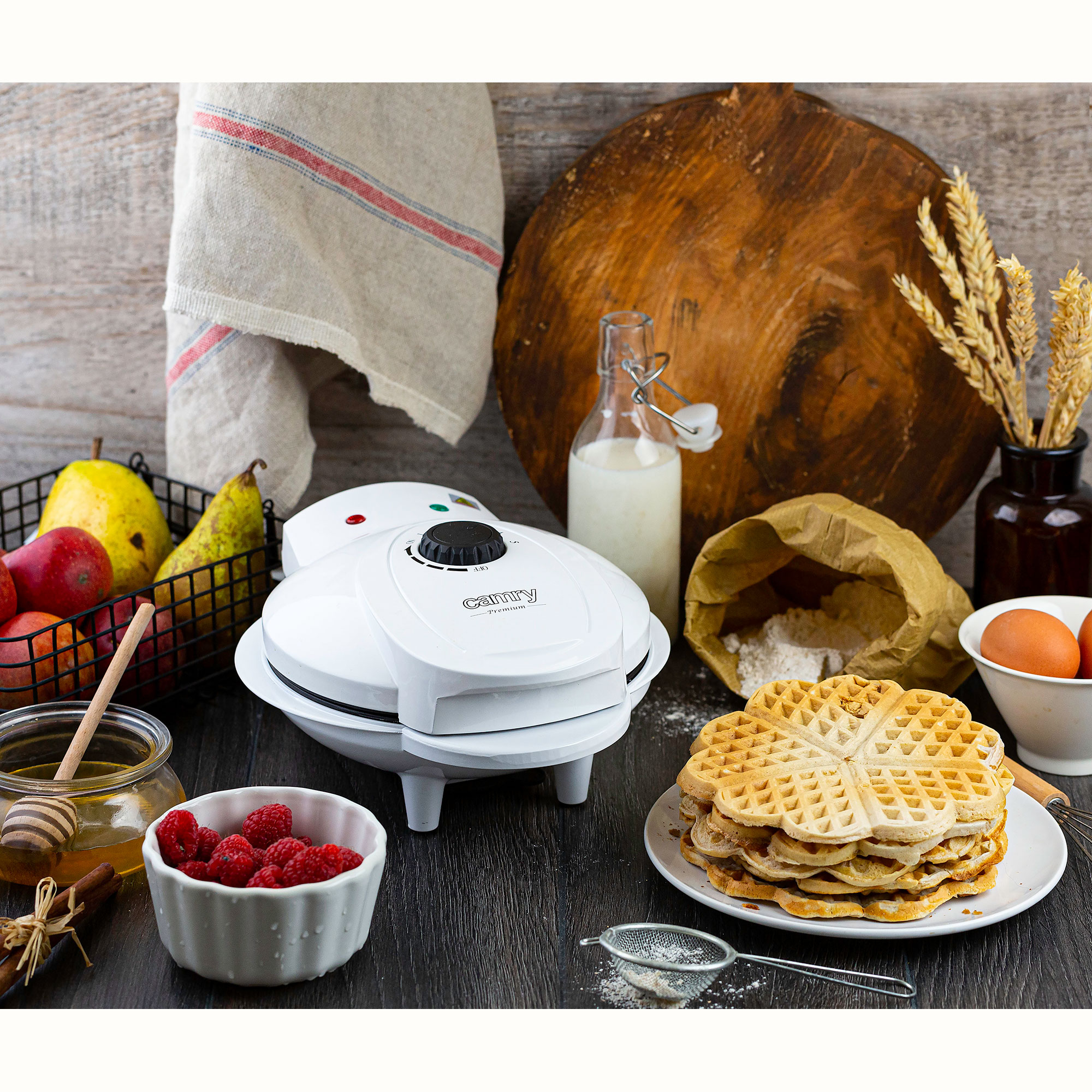 Una máquina de hacer gofres blanca con gofres, frutas, miel y leche en una mesa de madera.