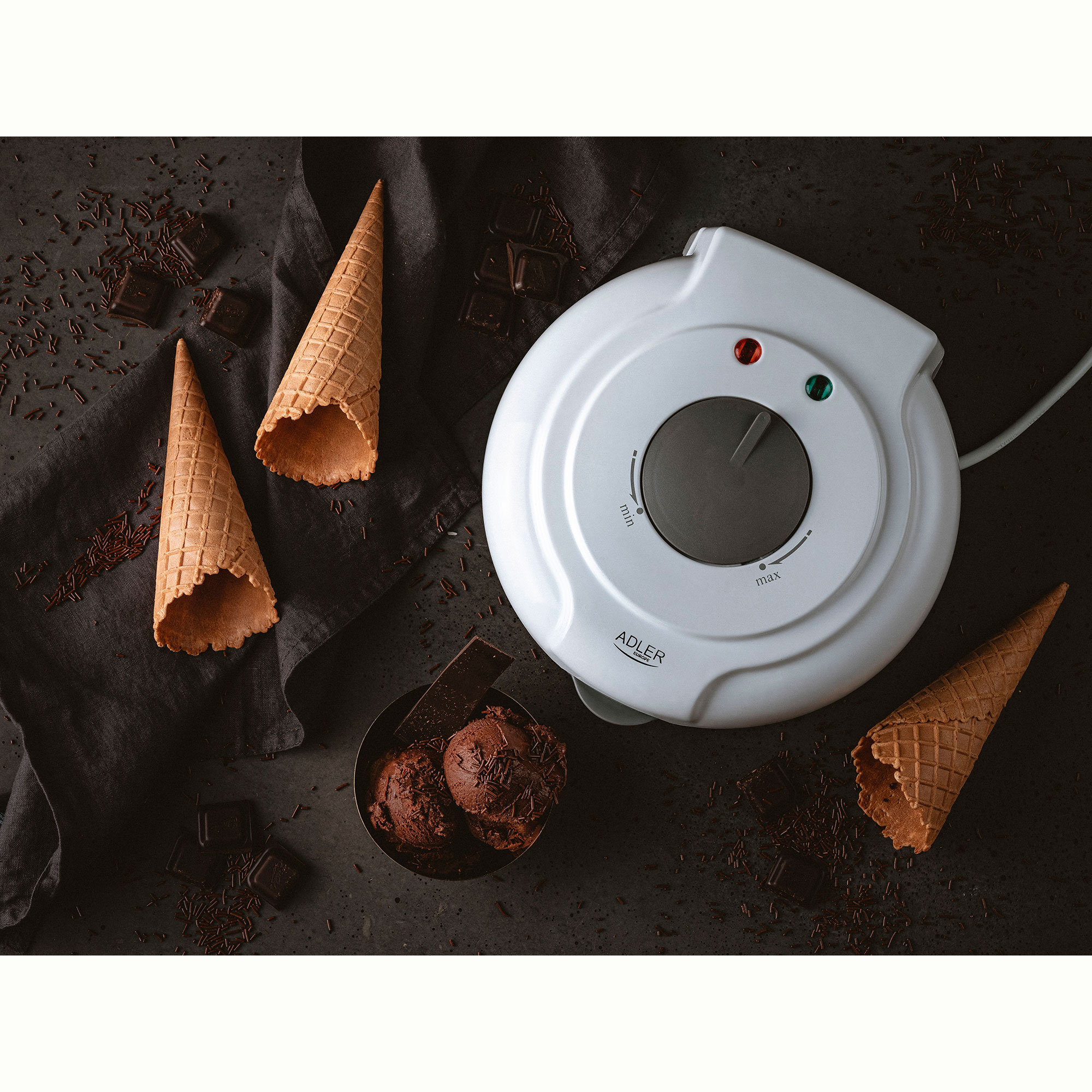 A waffle maker with cones, ice cream, and chocolate shavings on a dark surface. Overhead shot.