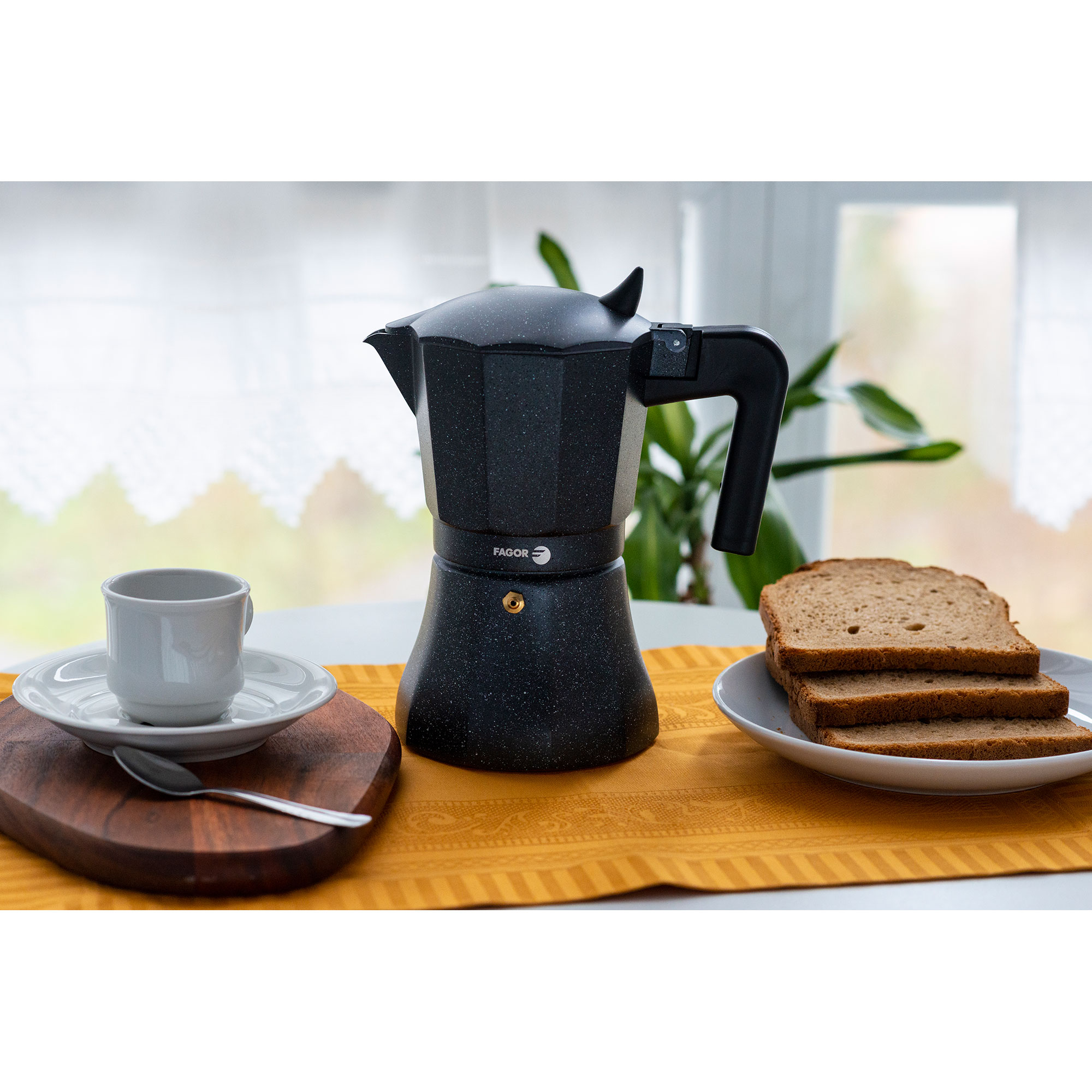 Cafetera moka negra con café, taza y rebanadas de pan sobre mantel amarillo.