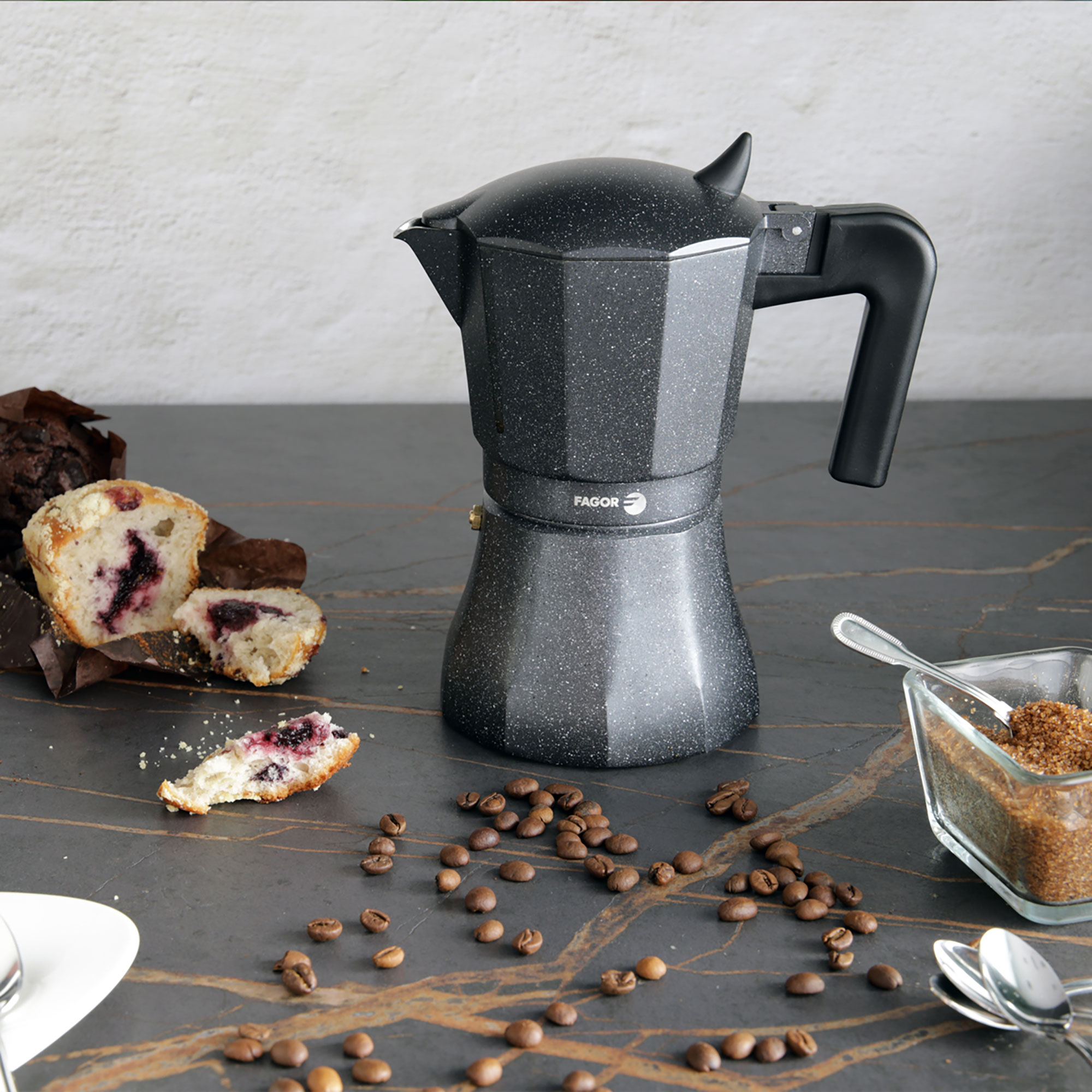 Cafetera espresso gris oscuro con granos de café, muffins y azúcar sobre una superficie oscura.