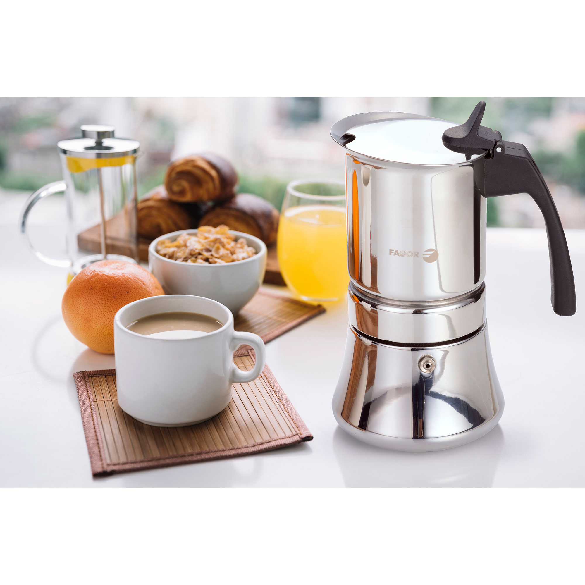 Una cafetera espresso Fagor de acero inoxidable, junto a una taza de café, fruta y pasteles, sobre una superficie blanca.