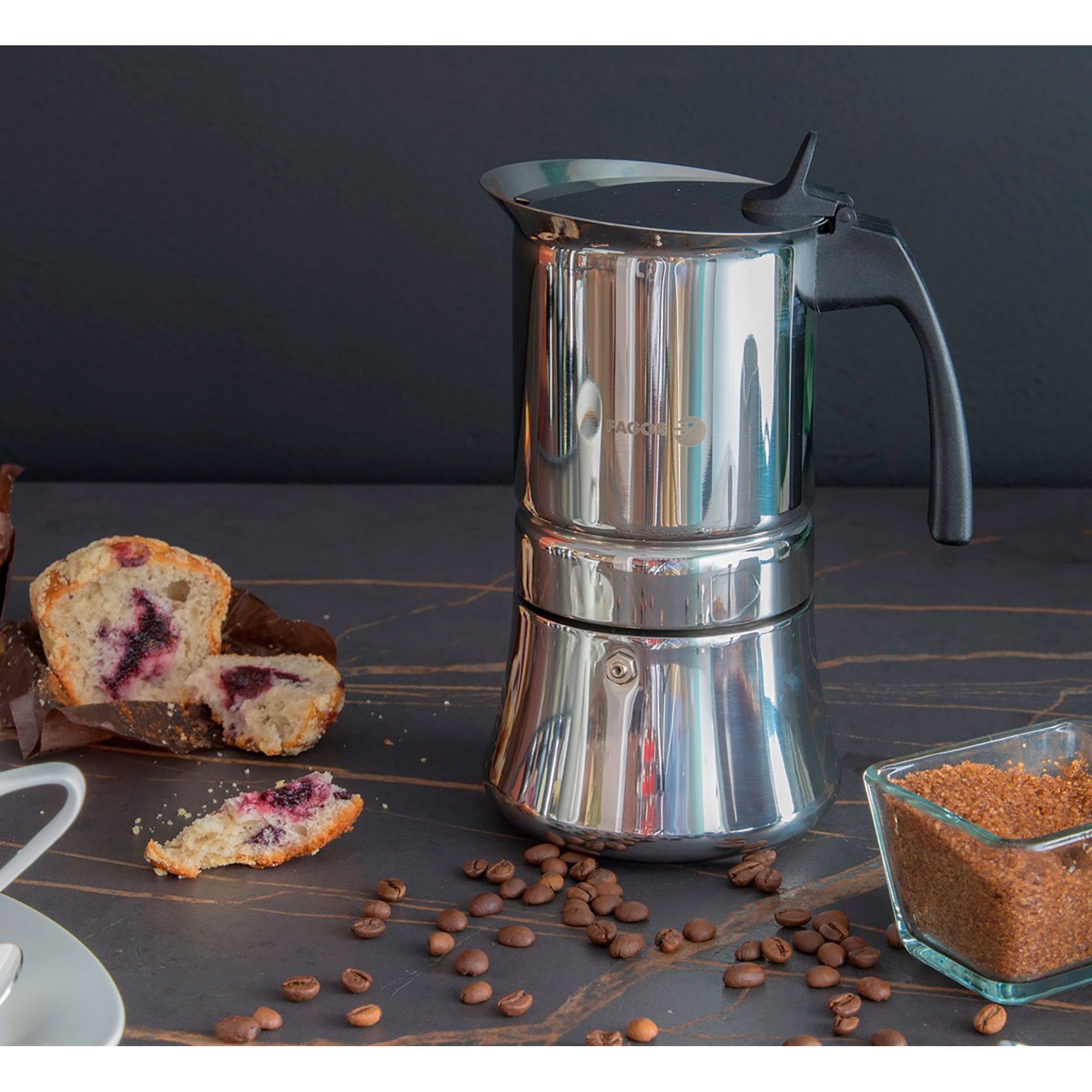 Cafetera italiana de acero inoxidable con muffins y granos de café sobre una superficie de mármol. El fondo es una pared oscura.