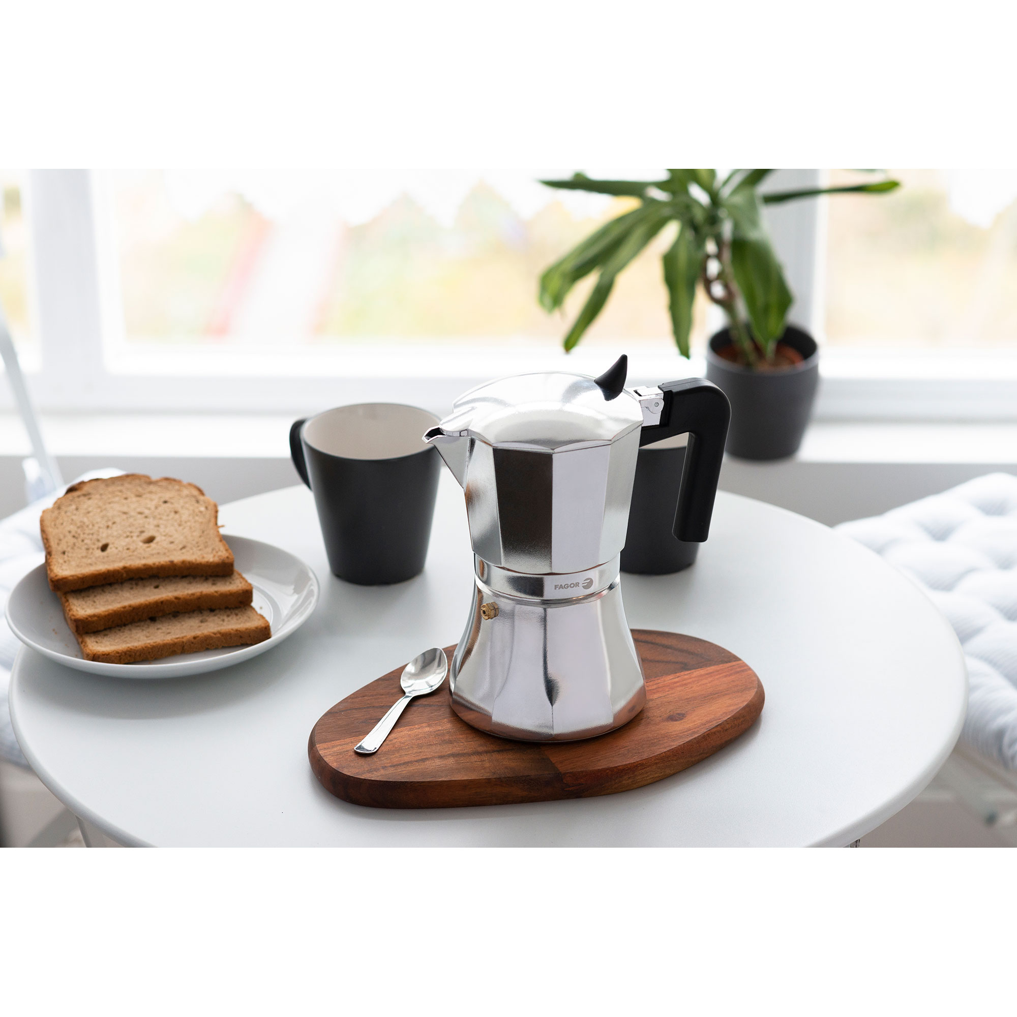Caffettiera espresso argentata su tagliere in legno, accanto a toast e tazza nera.