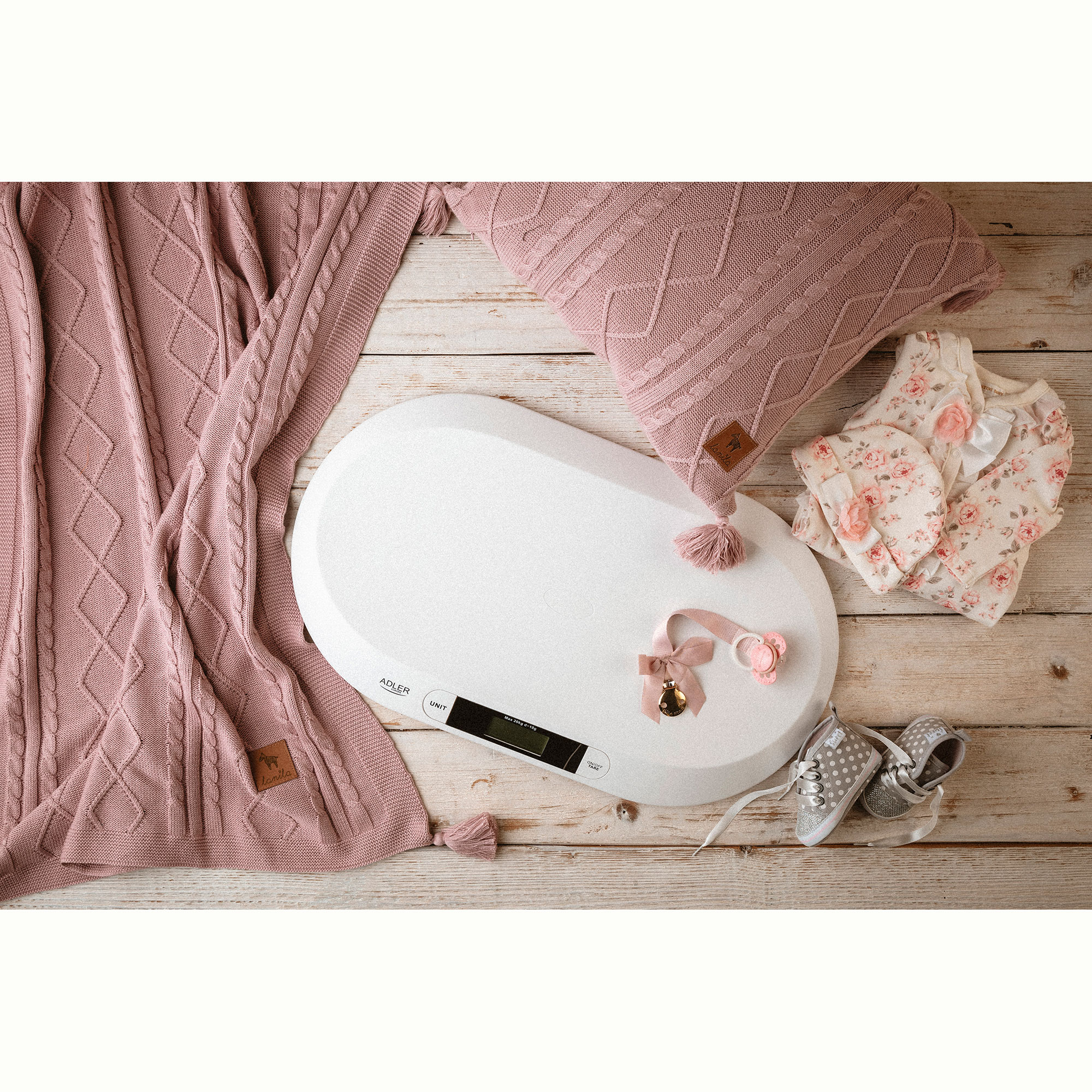 Balanza para bebés con manta, almohada, ropa y zapatos sobre una superficie de madera. Artículos rosas y blancos.