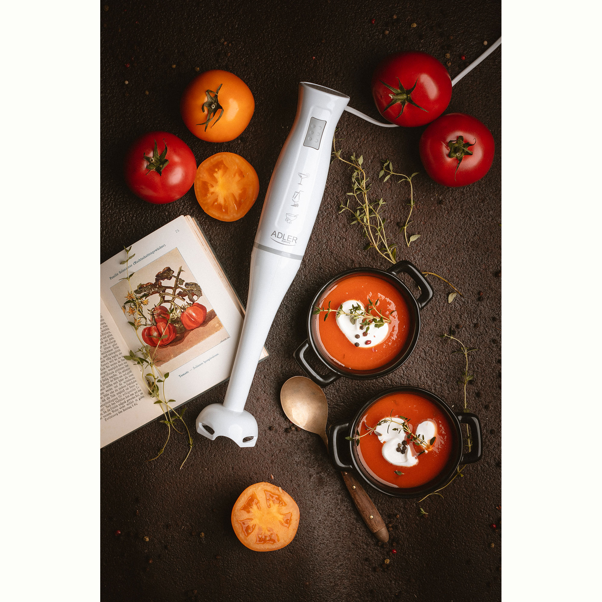 Een witte staafmixer met tomaten, soepkommen, tijm en een boek op een donker oppervlak.