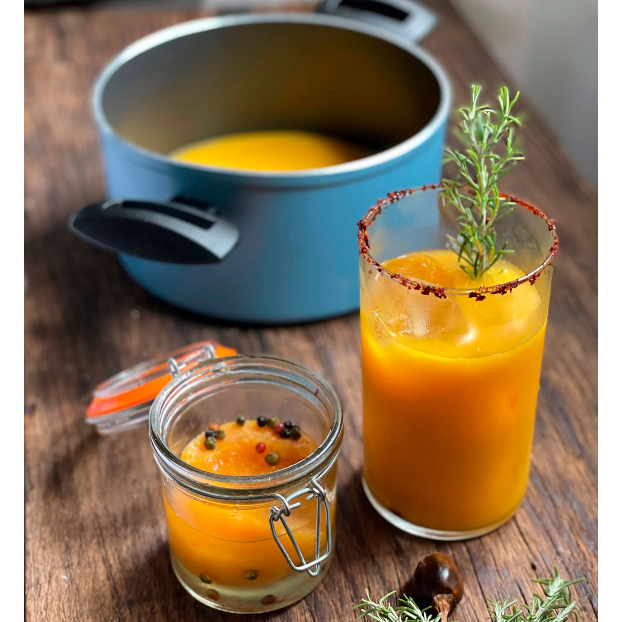 Bebida y sopa de naranja, romero en vaso, frasco con granos de pimienta y olla sobre madera.