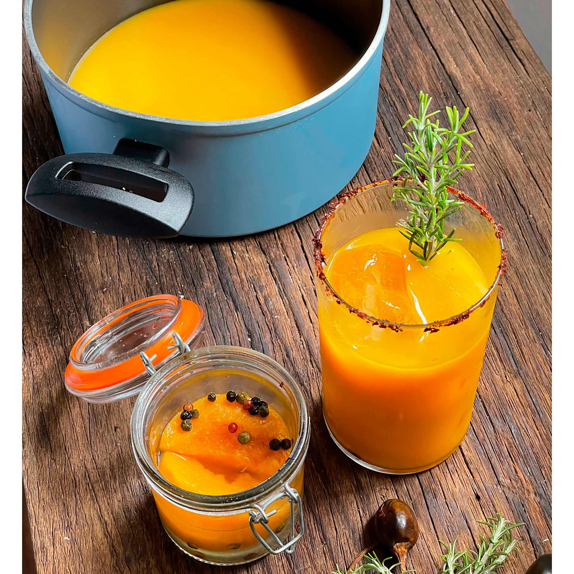 Mesa con sopa de naranja en olla, vaso y tarro, adornada con hierbas.