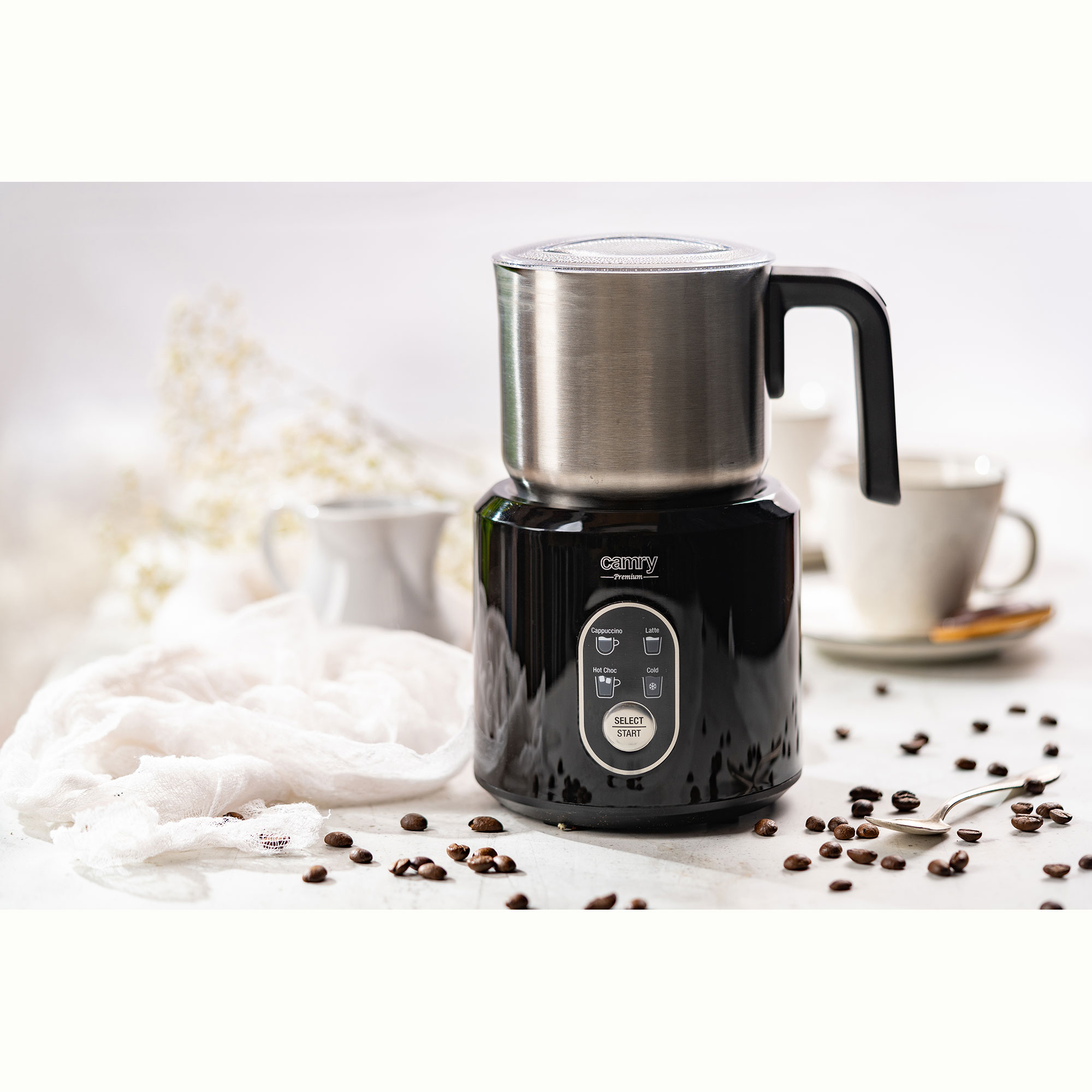 Espumador de leche negro y plateado con granos de café, tazas y cuchara sobre superficie blanca.