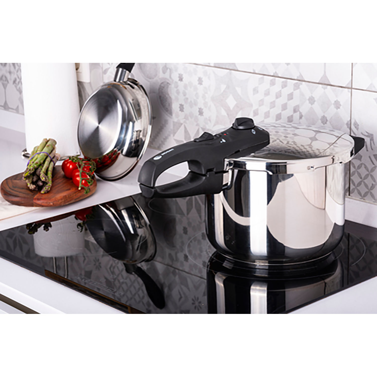 Olla a presión de acero inoxidable en una vitrocerámica negra. Espárragos, tomates y una sartén en la encimera.