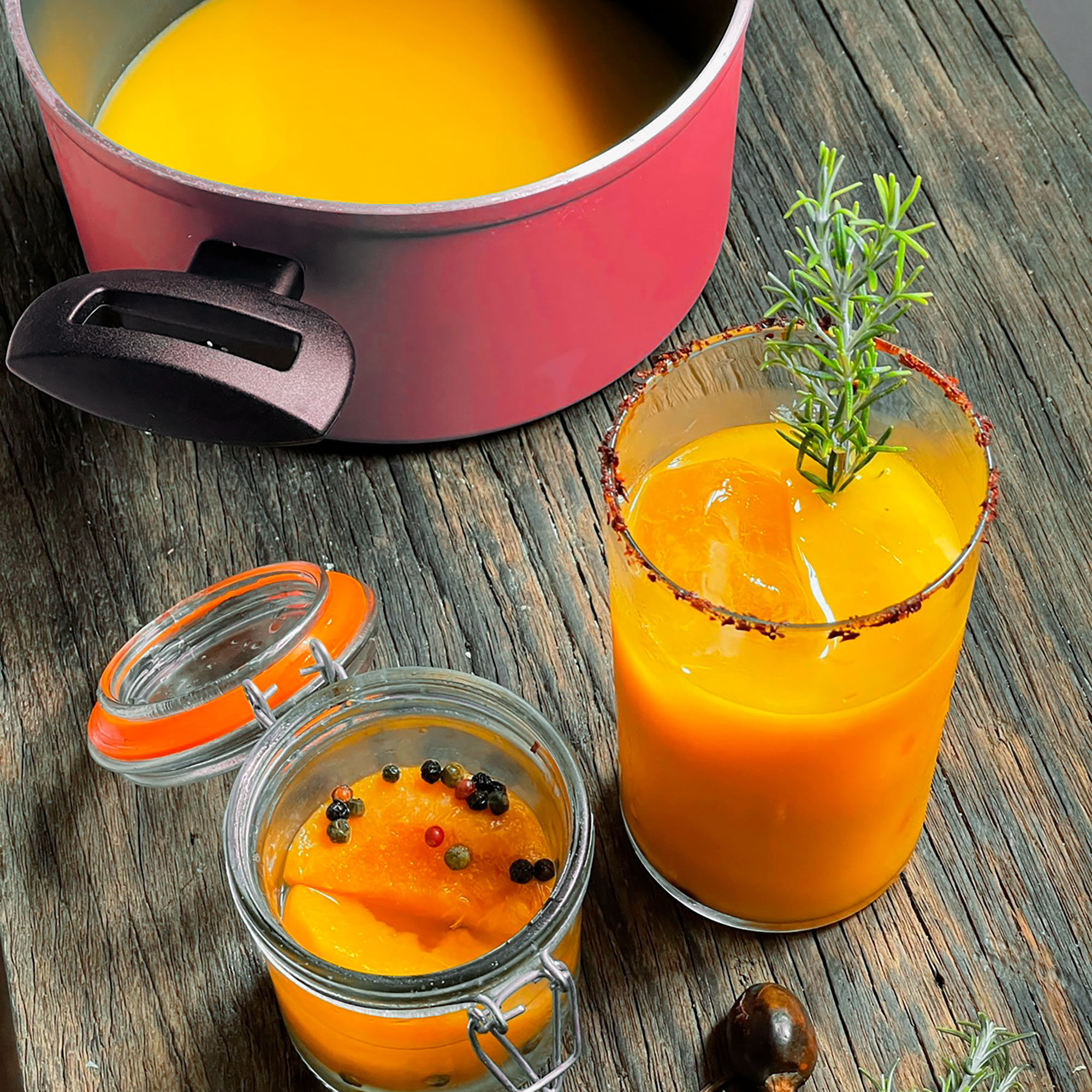 Vaso de bebida naranja con hielo y romero. Olla y tarro con ingredientes cerca. Madera.