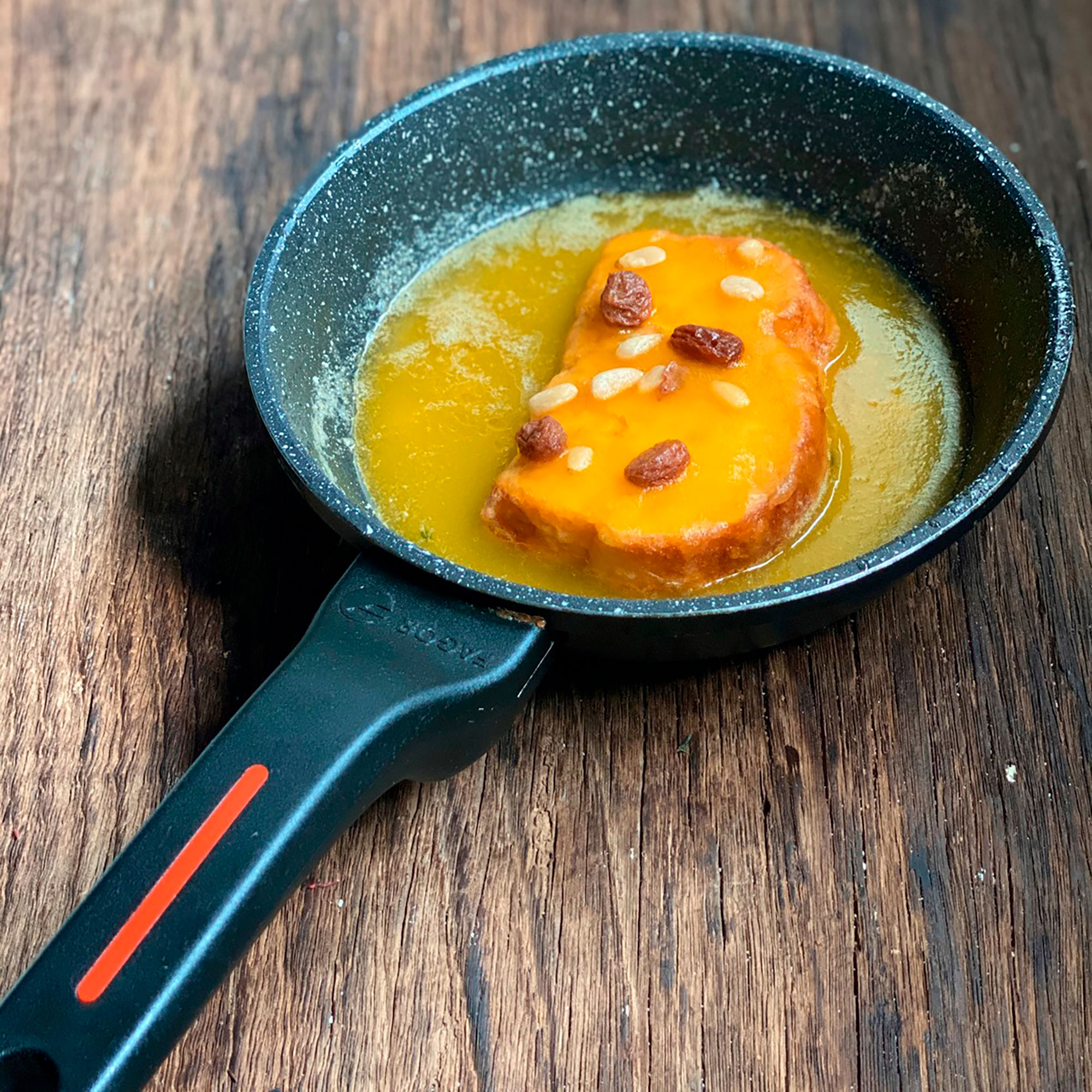 Pan frito en sartén. Salsa amarilla con pasas y piñones sobre mesa de madera.
