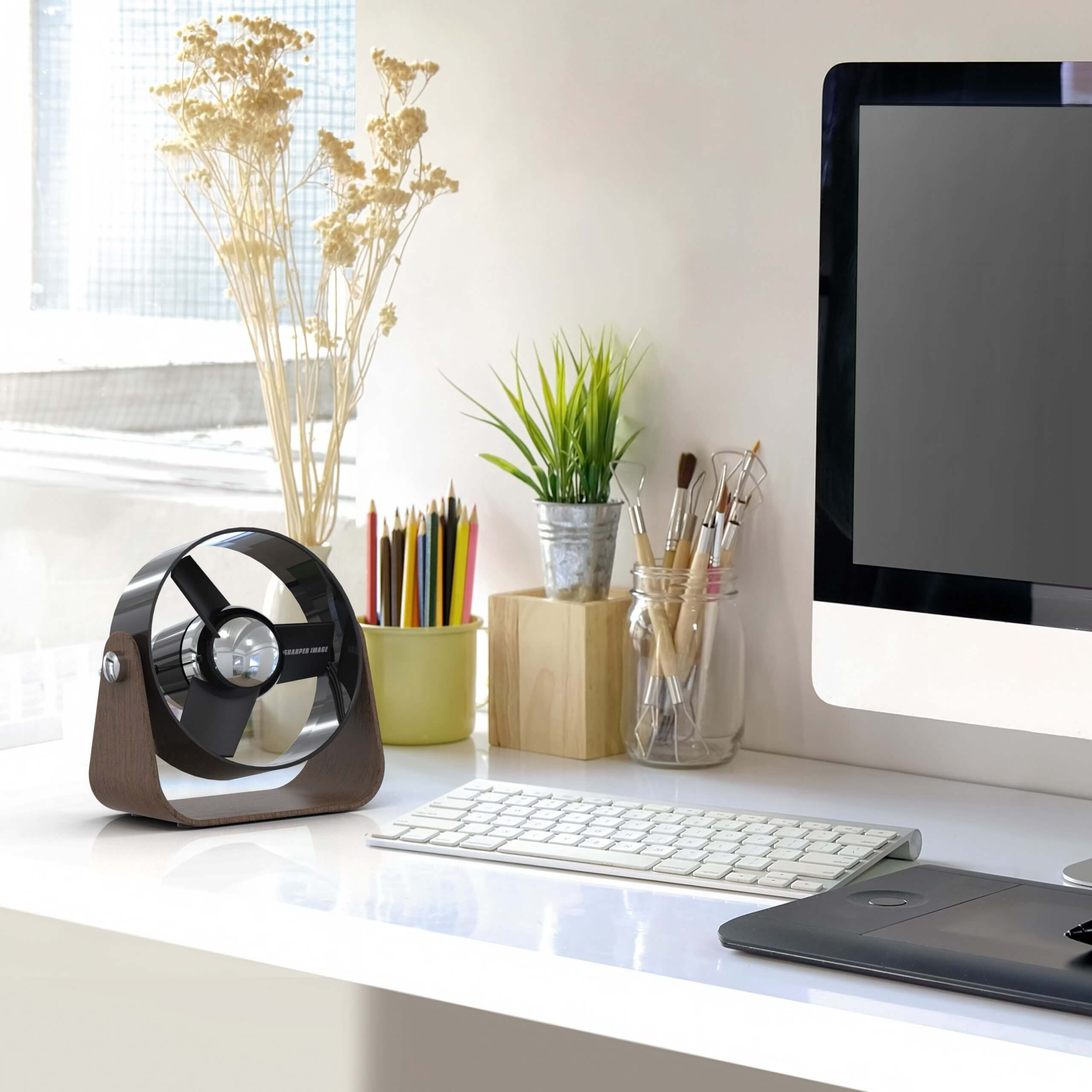 Schreibtisch mit Ventilator, Bleistiften, kleiner Pflanze, Tastatur und Monitor.