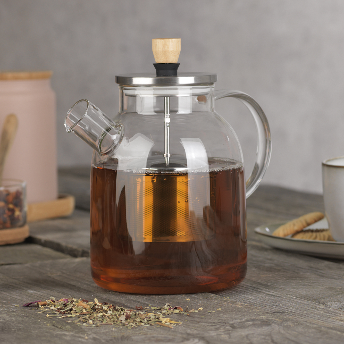 Gläserne Teekanne mit Tee gefüllt, mit Teefilter. Getrocknete Kräuter im Vordergrund.