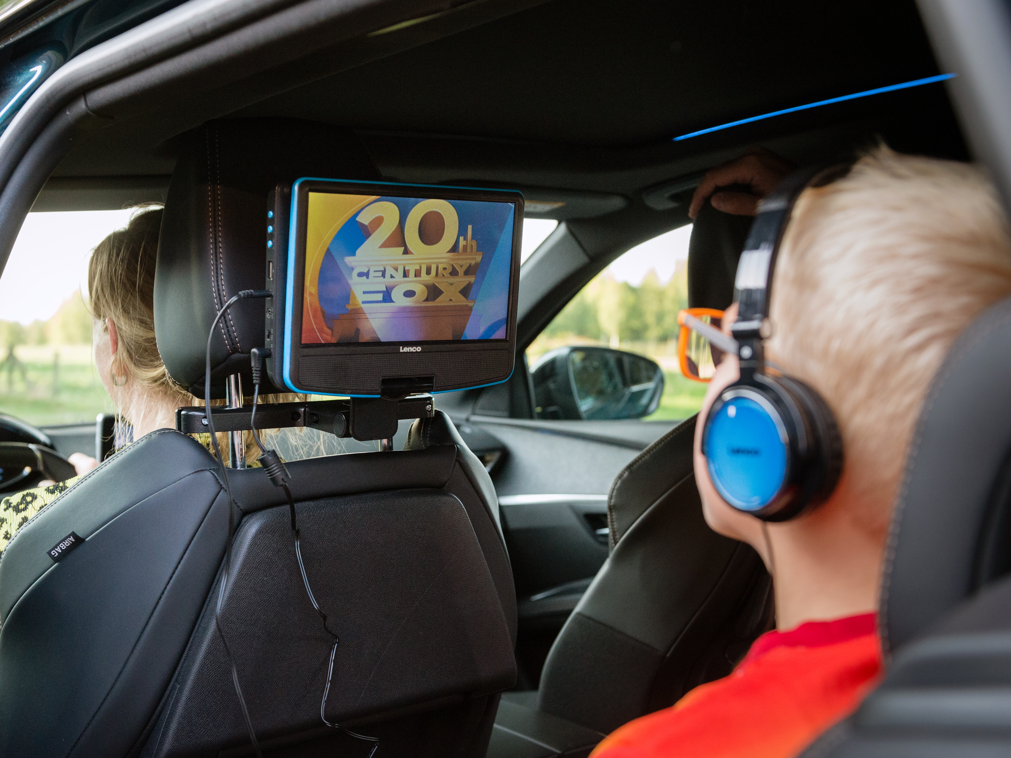 W samochodzie chłopiec ogląda 20th Century Fox na ekranie, w słuchawkach. Kobieta prowadzi.
