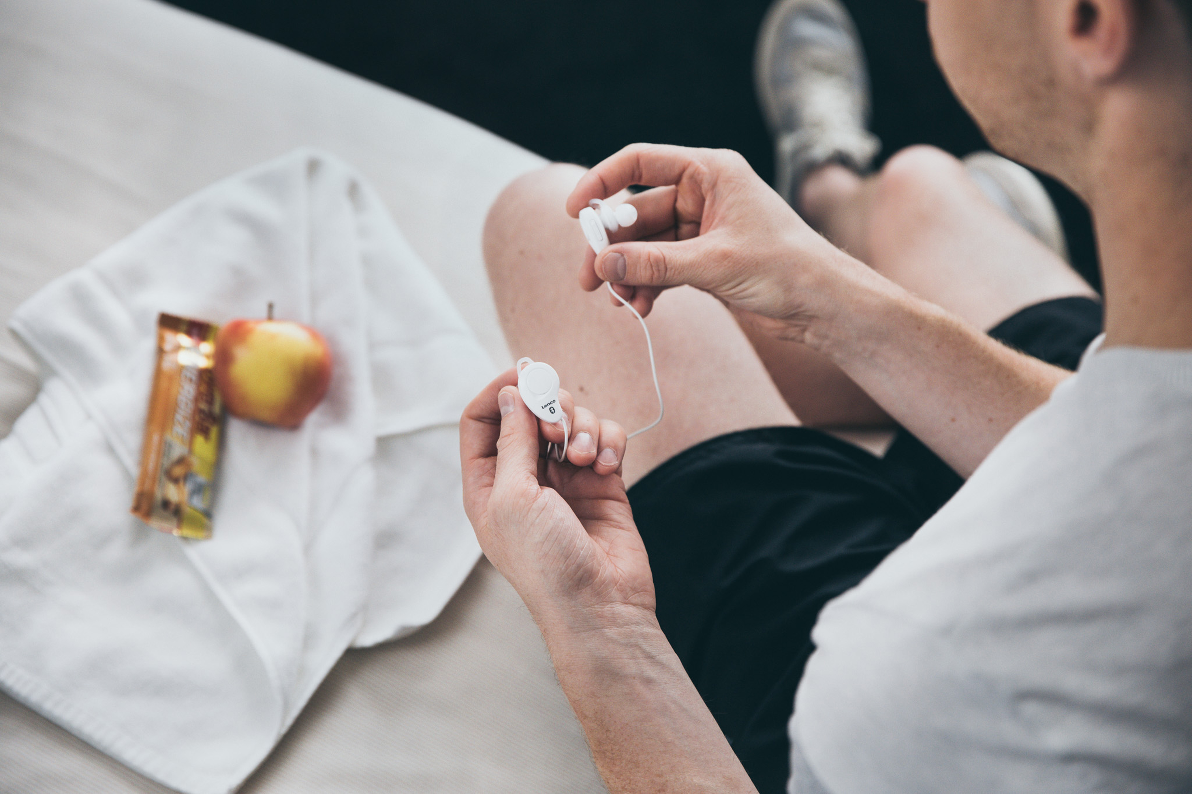 Eine Person hält Kopfhörer neben einem Apfel und einem Proteinriegel auf einem weißen Handtuch.