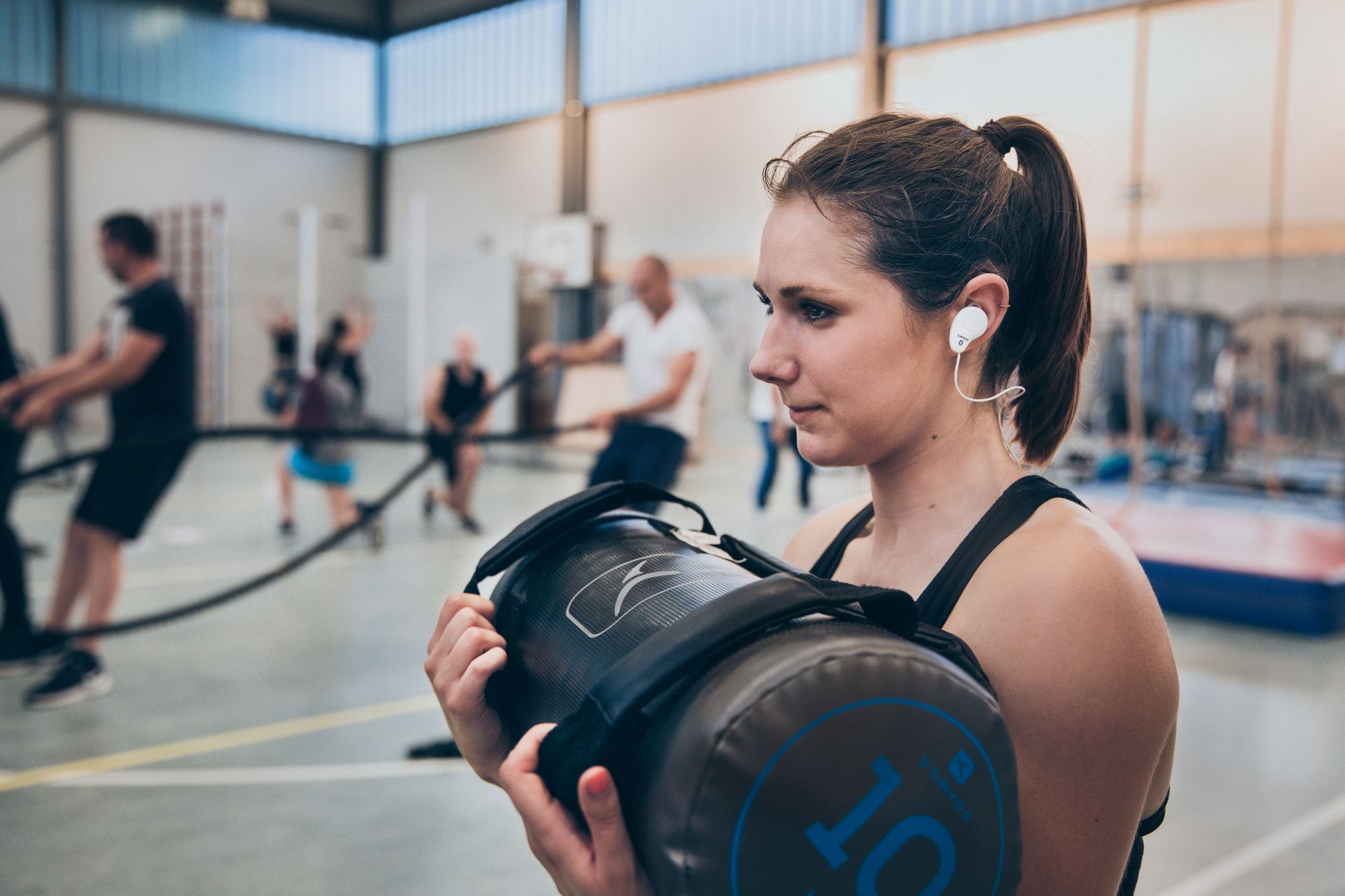 Eine Frau im Fitnessstudio hält ein schwarzes Gewicht, andere Personen im Hintergrund.