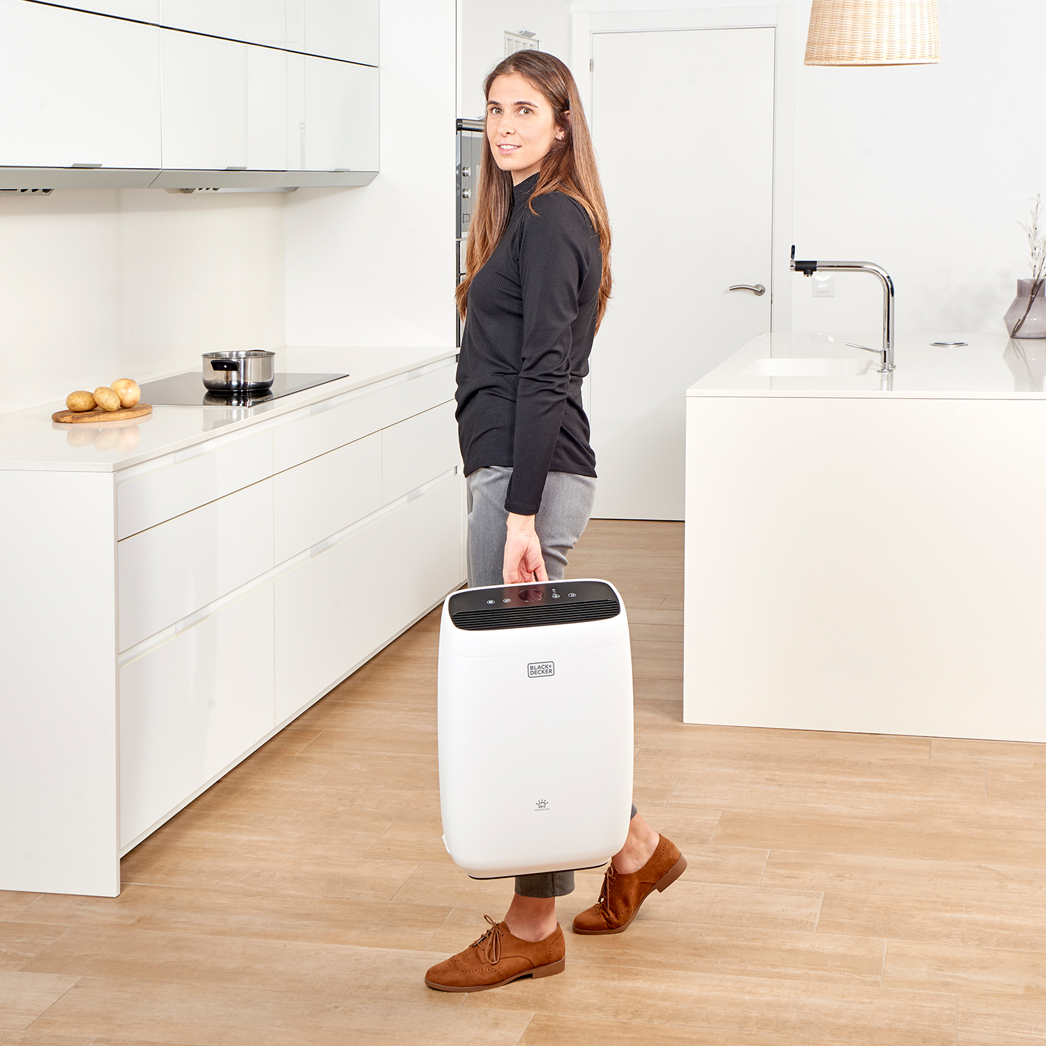 Donna in nero tiene purificatore d'aria bianco in cucina. Pavimento in legno, armadi bianchi ed elettrodomestici visibili.