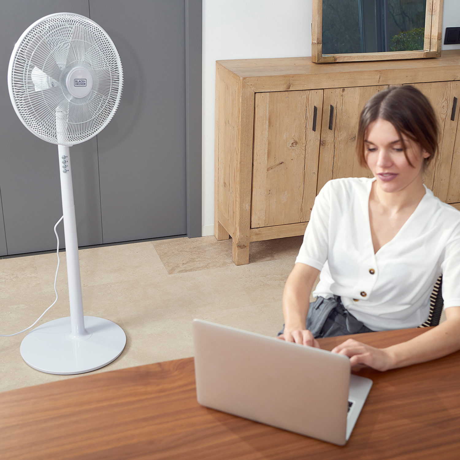 Kobieta z laptopem siedzi obok białego wentylatora. Za nią jest drewniana szafka.