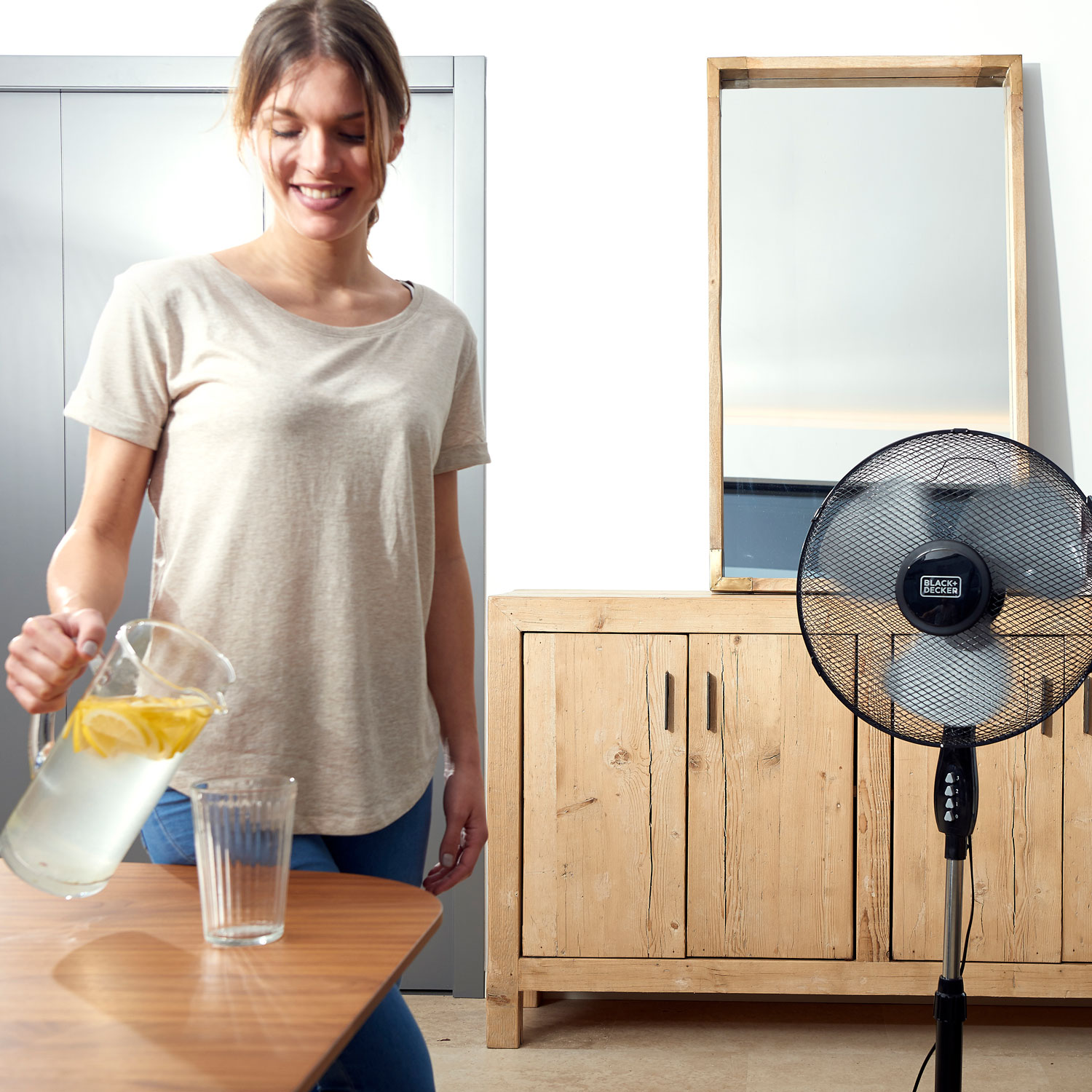 Kobieta nalewa wodę z cytryną z dzbanka do szklanki, w tle wentylator i szafka.