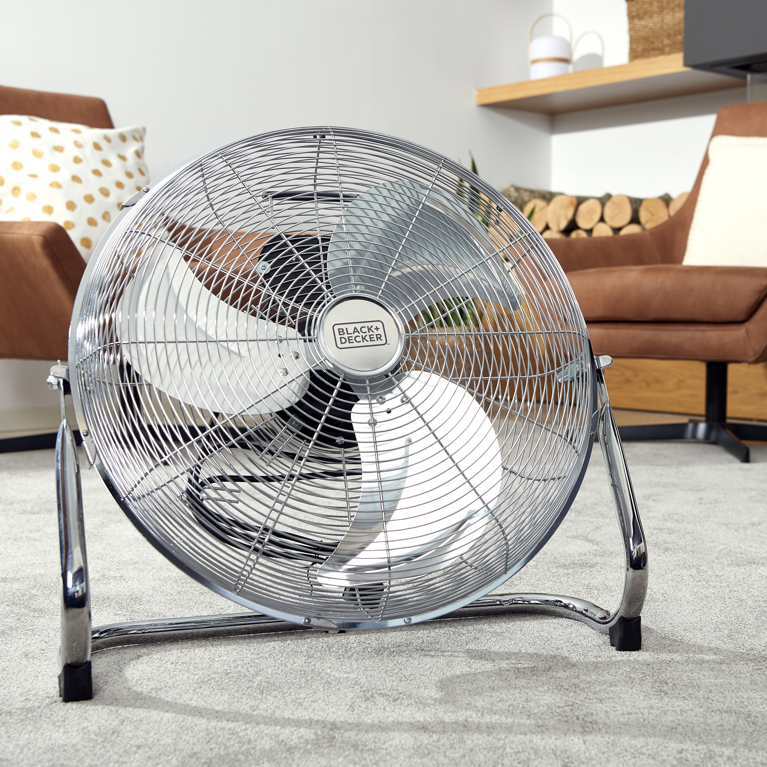 Un ventilatore in metallo con tre pale su un pavimento grigio rivestito di moquette.